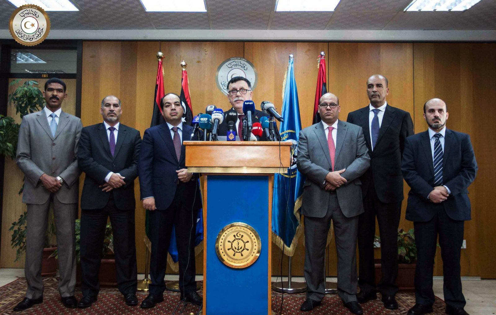 The image shows a group of eight men standing in a formal setting, likely a press conference or official announcement. There is a podium at the center with a seal or emblem, and the speaker appears to be addressing the audience. The backdrop features flags, suggesting a governmental or institutional context. The men are dressed in suits and ties, conveying a professional atmosphere.