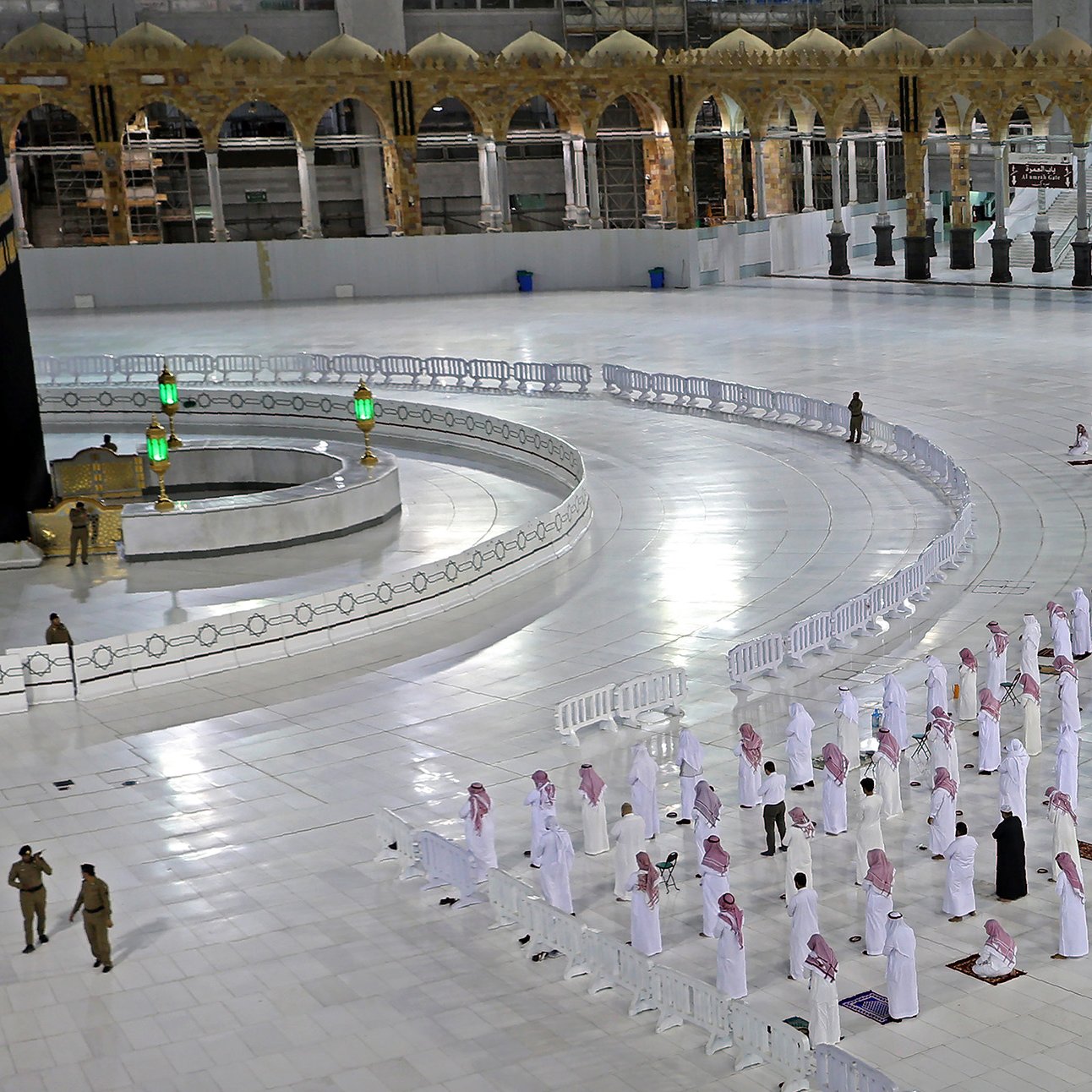 The image depicts a scene from the Grand Mosque in Mecca, known as the Masjid al-Haram. It features the Kaaba, a black cube-shaped structure covered in a black cloth with gold embroidery, which is the focal point of Islamic worship. Surrounding the Kaaba, there are congregational worshippers who appear to be lined up in rows, praying. The area is spacious and well-lit, with white marble flooring. In the background, there are architectural elements characteristic of the mosque, including arched structures. Security personnel can also be seen in the image, emphasizing the organized nature of the gathering.