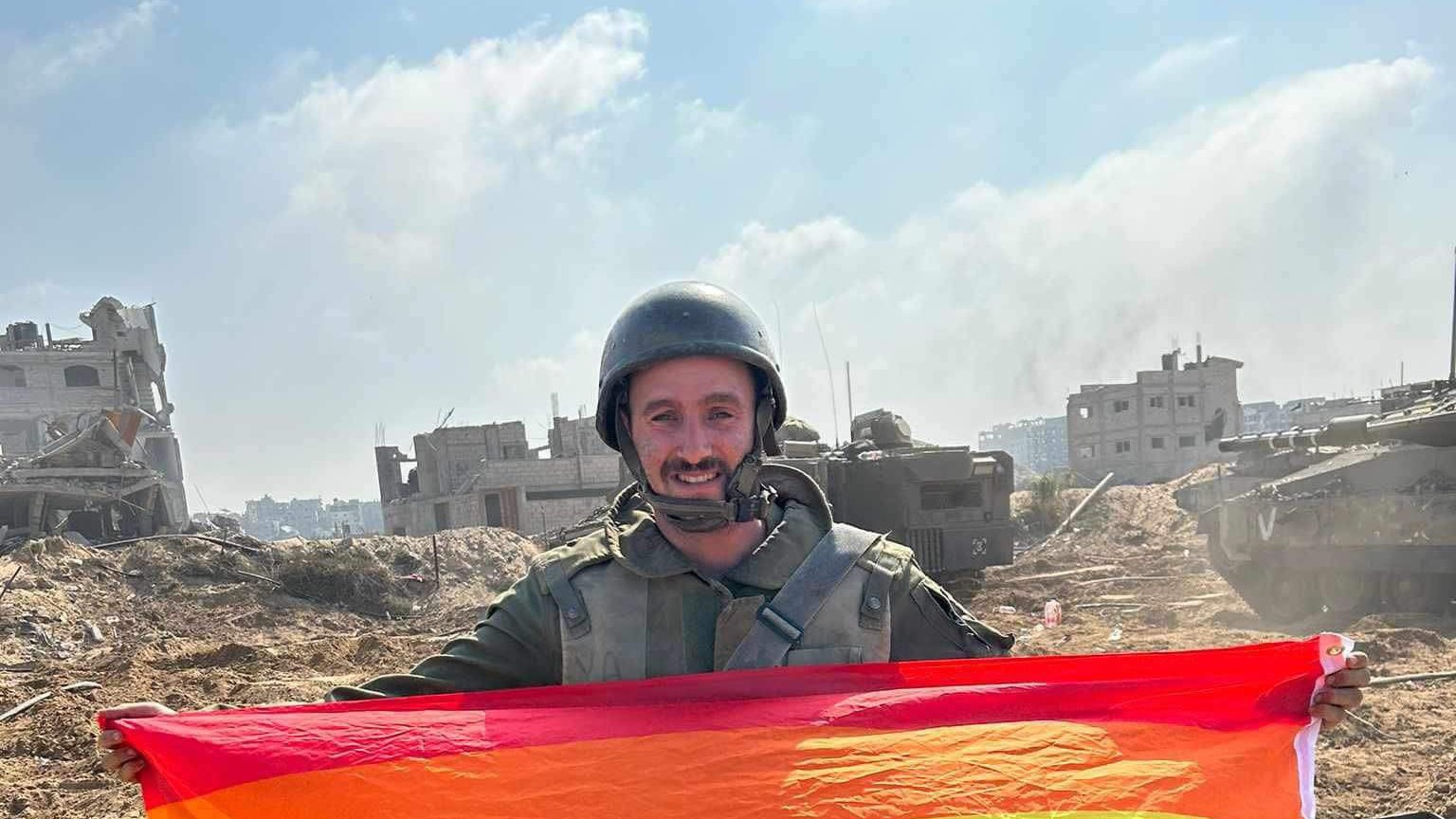 Un soldat souriant tient un drapeau arc-en-ciel sur un terrain dévasté.