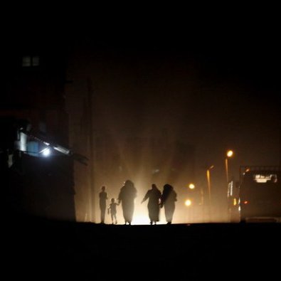 L'image montre un groupe de personnes marchant sur une route sombre, illuminée par des lumières vives au loin. Les silhouettes des individus sont visibles, créant une ambiance mystérieuse. En arrière-plan, on aperçoit des bâtiments et d'autres sources de lumière, suggérant un environnement urbain. L'atmosphère est à la fois sombre et intrigante, mettant en avant le contraste entre l'ombre et la lumière.