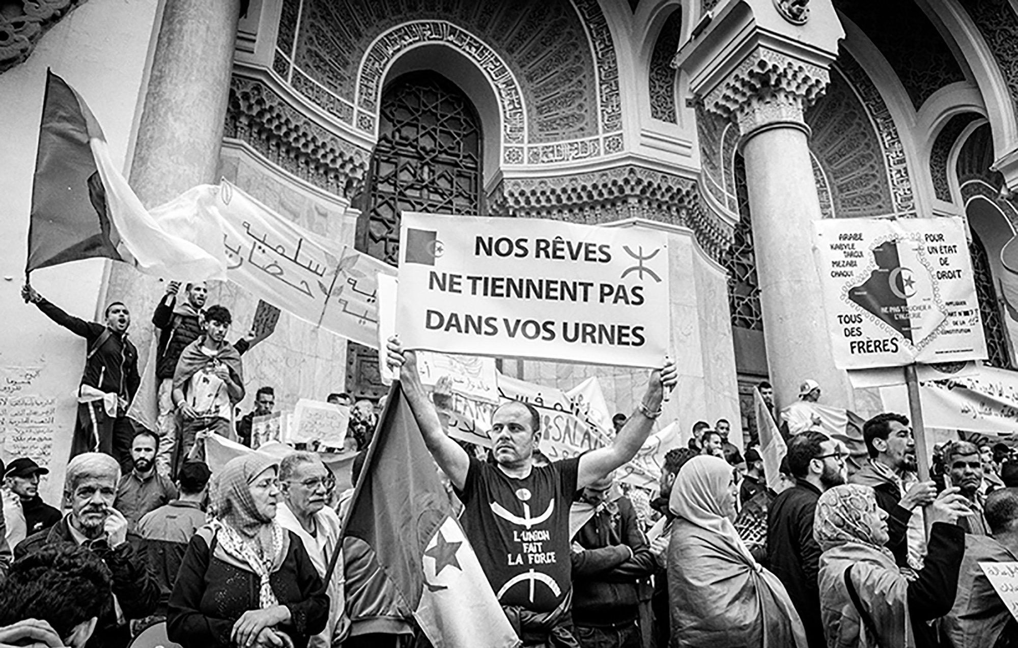 L'image montre une scène de manifestation où plusieurs personnes sont rassemblées. Au premier plan, un homme tient une grande pancarte qui affiche le message : "NOS RÊVES NE Tiennent PAS DANS VOS URNES". Les manifestants entourant cet homme expriment des sentiments de protestation, avec des drapeaux et des banderoles visibles. L'atmosphère semble chargée d'émotion et d'engagement politique, le tout dans un cadre architectural imposant. La photo est en noir et blanc, ce qui accentue l'intensité de la situation.