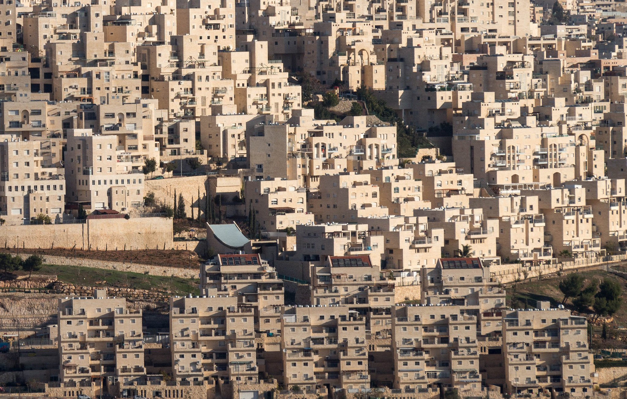 L'image présente un paysage urbain dense, constitué de nombreux bâtiments aux formes géométriques. Les structures sont principalement de couleur claire, avec des nuances de beige et de blanc. On peut observer des immeubles empilés les uns sur les autres, créant une apparence presque uniforme. Le terrain autour des bâtiments semble légèrement vallonné, avec des zones de végétation sporadique. Il y a également des éléments architecturaux distincts, tels qu'un toit qui se distingue parmi l'ensemble des constructions. L'ensemble donne une impression de modernité et d'urbanisation.