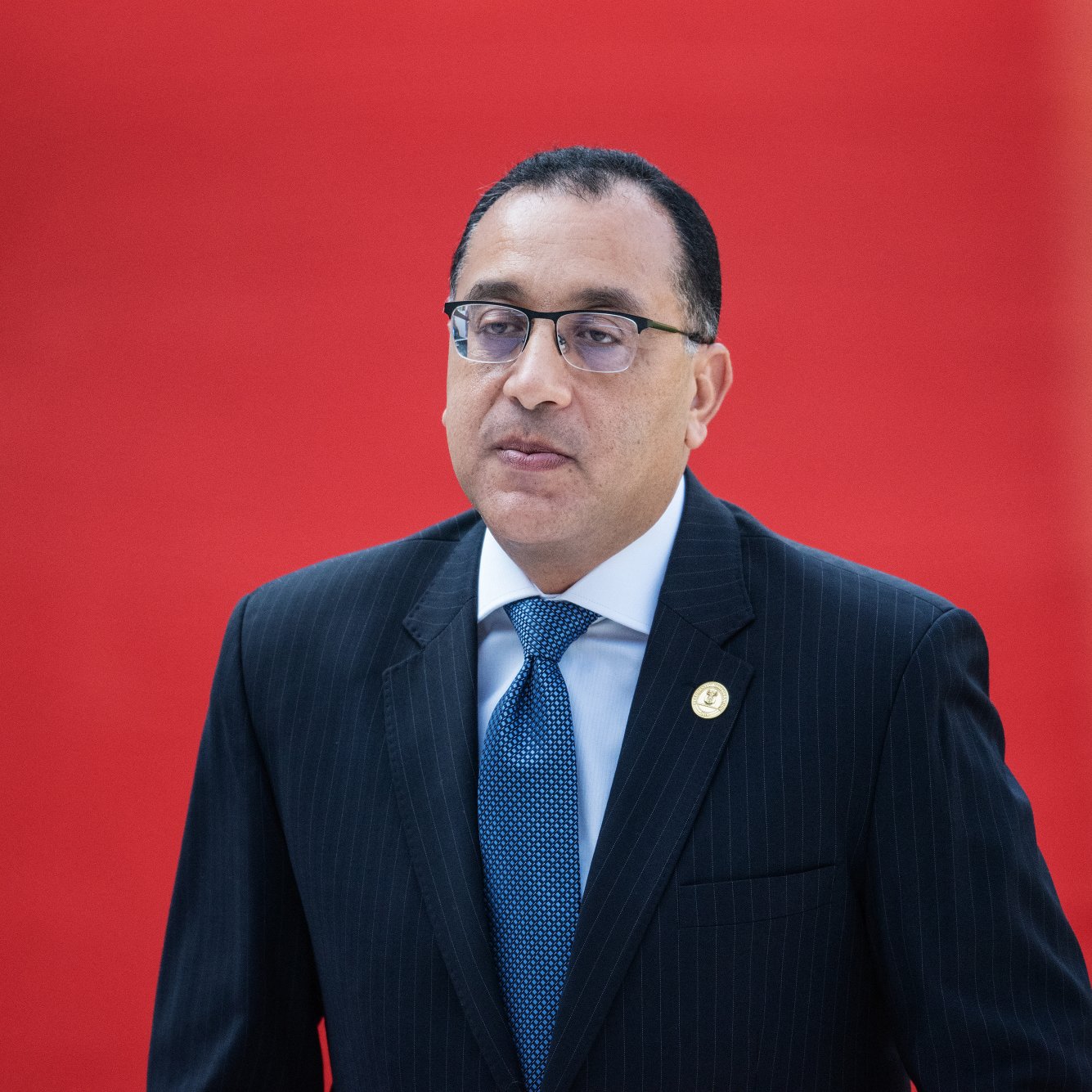 L'image montre un homme en costume noir avec une chemise blanche et une cravate bleue. Il porte des lunettes et a une expression sérieuse. L'arrière-plan est rouge, ce qui crée un contraste vif avec sa tenue. L'homme semble être dans un cadre officiel ou lors d'un événement important.