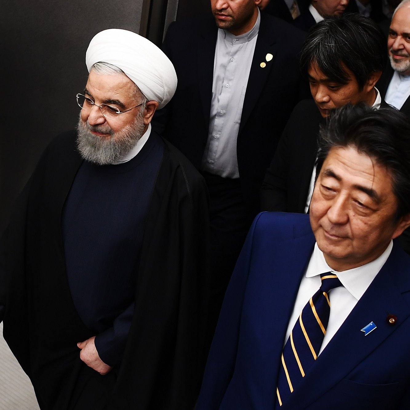 L'image montre deux hommes marchant côte à côte dans un environnement formel. L'un d'eux porte une tenue sombre avec une coiffure distinctive et un turban blanc, tandis que l'autre est habillé de manière formelle en costume. Ils semblent être entourés de plusieurs autres personnes habillées aussi de manière professionnelle. L'atmosphère dégage un air de réunion ou d'événement officiel.