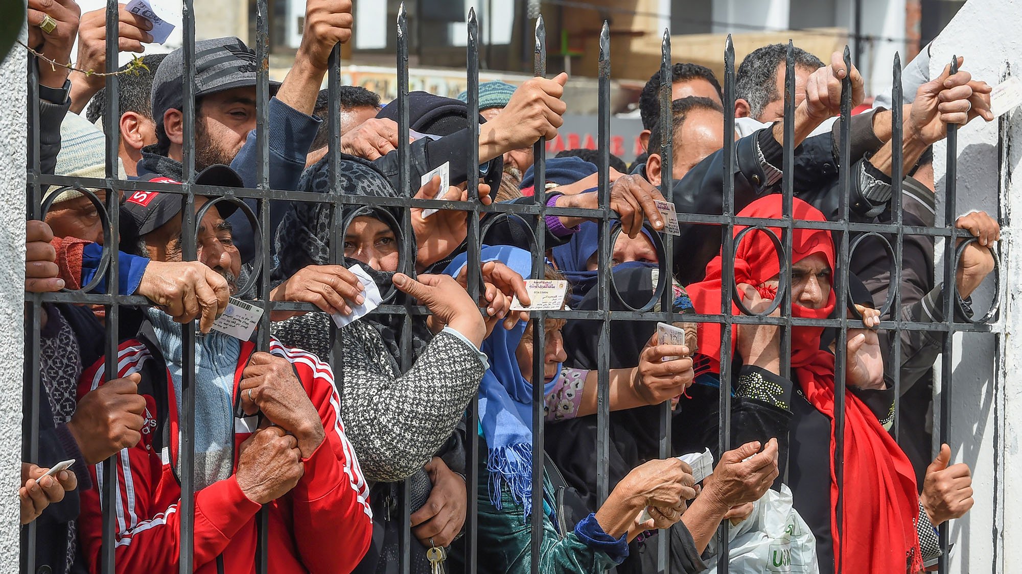 تظهر في الصورة مجموعة من الأشخاص المتجمعين خلف سياج حديدي. يرفع البعض منهم أوراقًا أو بطاقات، بينما يبدو التركيز على حركتهم وتصرفاتهم، مما يوحي بأنهم قد يكونون في انتظار شيء ما أو يحاولون الوصول إلى جهة معينة. الأجواء تعكس حالة من الترقب أو الأمل، مع تنوع في الملابس، مما يشير إلى خلفيات مختلفة.