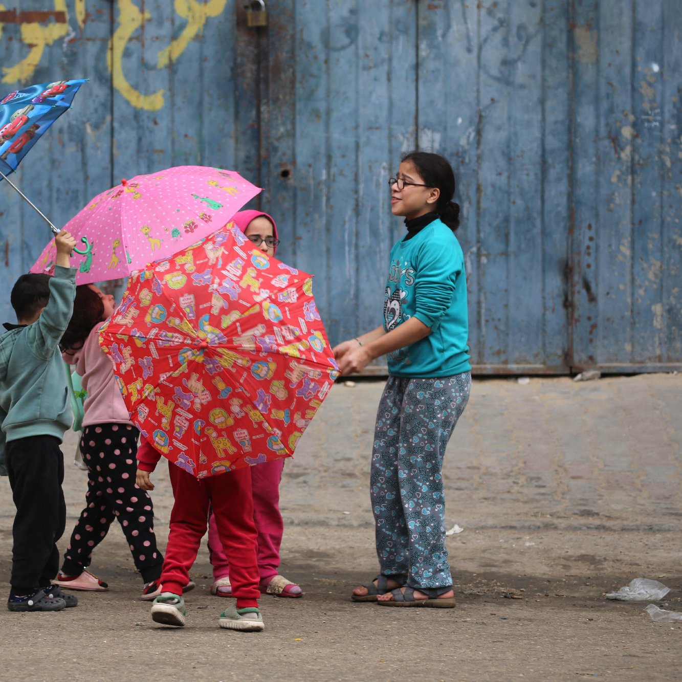 در تصویر، چند کودک در حال بازی با چترهای رنگارنگ هستند. چترها طرح‌های شاد و جذابی دارند و کودکان در حالی که در دایره‌ای ایستاده‌اند، به هم نزدیک هستند. زمینه تصویر یک دیوار آبی رنگ و زمین سنگ‌فرش شده است.