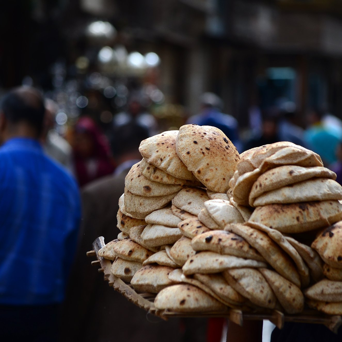 L'image montre un vendeur de pain dans une rue animée. Il porte une grande corbeille chargée d'un tas de pains ronds et dorés, souvent appelés "pita" ou "naan". En arrière-plan, on aperçoit des silhouettes de personnes se déplaçant dans la rue, créant une atmosphère vivante et dynamique. L'environnement semble plutôt urbain, avec des bâtiments et d'autres vendeurs visibles au loin.