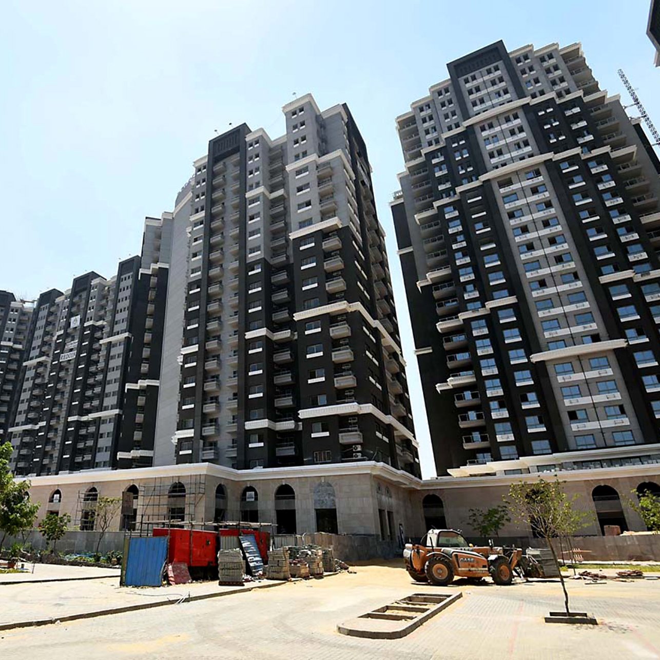 تظهر في الصورة مجموعة من المباني السكنية الحديثة والمرتبة. تتميز العمارات بتصميمها المعماري العصري، حيث تحتوي على واجهات بلونين رئيسيين: الأسود والأبيض. تمتاز البناية بارتفاعها وكثرتها، ويبدو أن هناك أعمال بناء وإصلاحات جارية في المنطقة المحيطة. كما توجد بعض الأشجار والنباتات، مما يضفي لمسة خضراء على المشهد. السماء تبدو صافية ومشرقة، مما يعطي شعورًا بالزمن الجميل.