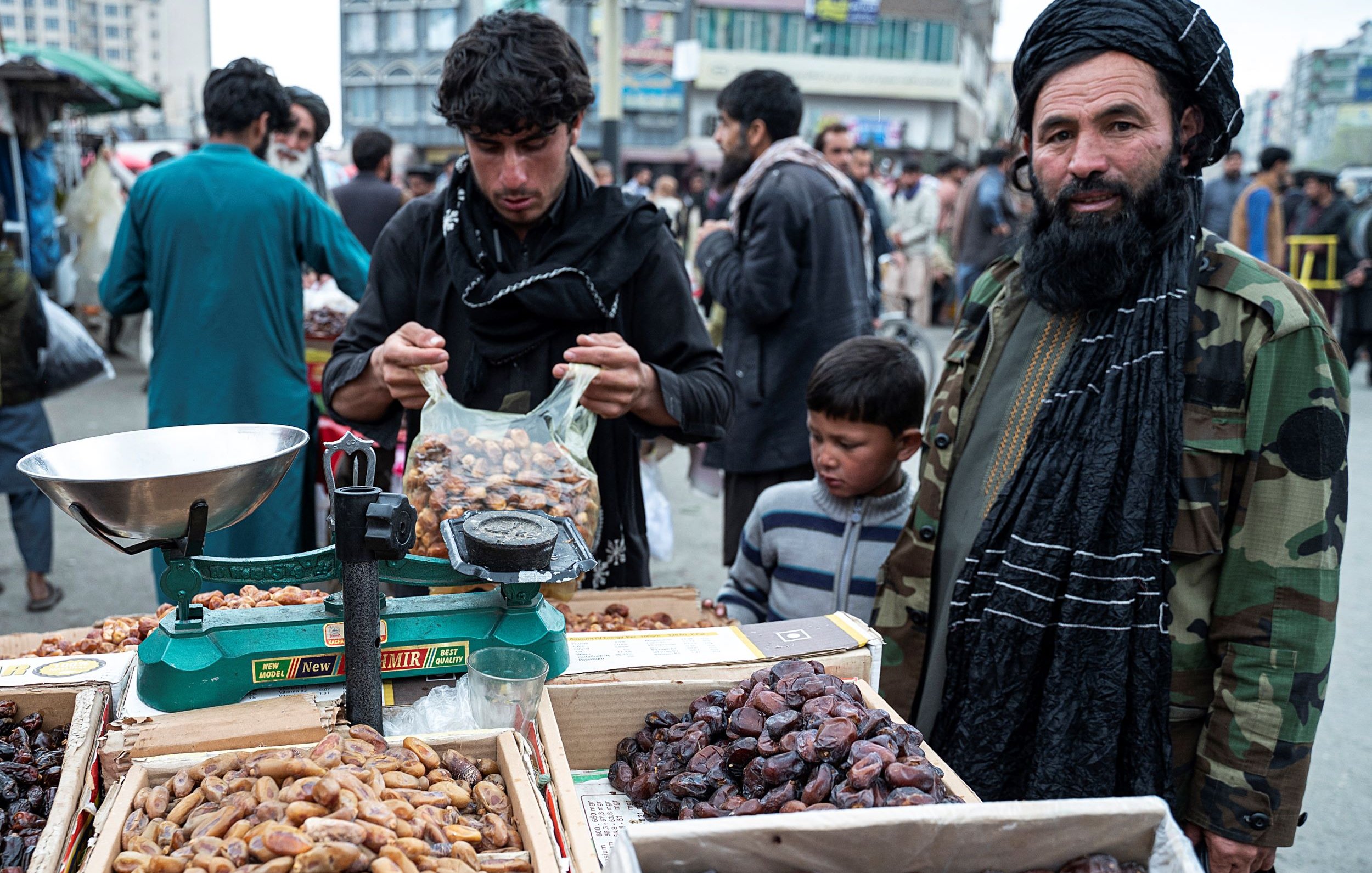 L'image montre un marché en plein air, probablement dans un pays asiatique. Au premier plan, on voit un homme portant un turban et une barbe, habillé d'une veste militaire. À côté de lui, un enfant observe. L'homme a l'air de s'occuper d'une balance et de dates, qui sont présentées dans des caisses en bois à ses côtés. En arrière-plan, on aperçoit d'autres personnes, ce qui donne une impression de foule animée et d'activité commerciale. L'environnement semble rural et traditionnel, avec des bâtiments flous au fond.