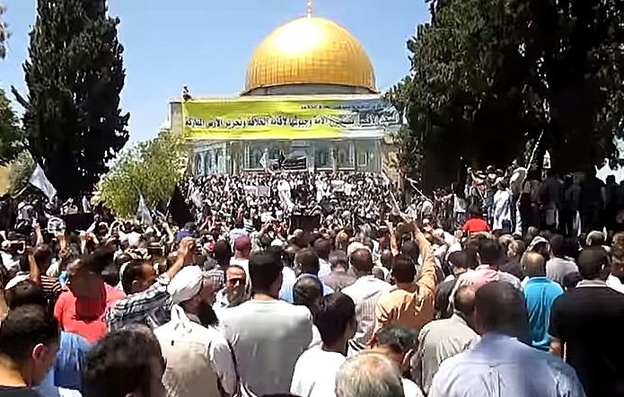 L'image montre une foule rassemblée devant un bâtiment orné d'un dôme doré. Les participants semblent être engagés dans une manifestation ou un rassemblement, avec des drapeaux et des pancartes visibles. Les arbres environnants ajoutent au décor, et l'atmosphère générale paraît dynamique, marquée par une forte présence de personnes.