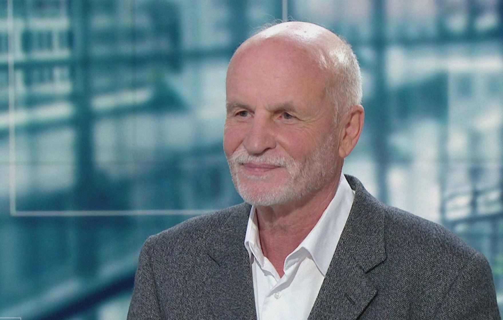 Homme âgé avec cheveux gris, souriant, portant un costume sobre sur fond flou.