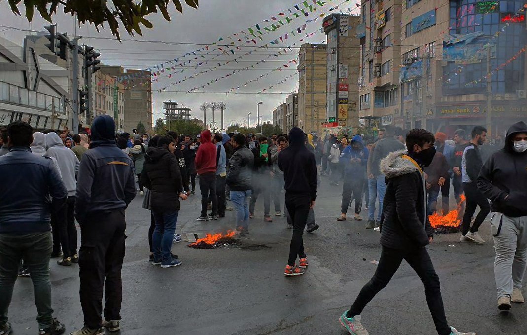 تظهر الصورة مجموعة كبيرة من الناس في منطقة حضرية، حيث يبدو أن هناك تظاهرة أو تجمع حاشد. يتواجد المتظاهرون في الشارع، وبعضهم يرتدي أقنعة وجه. هناك دخان يتصاعد من نيران صغيرة مشتعلة على الأرض. السماء تبدو غائمة، وهناك زينة ملونة معلقة في الجو، مما يشير إلى وجود نشاطات أو احتفالات بجانب المظاهرات. الأجواء تعكس التوتر نتيجة الظاهر.