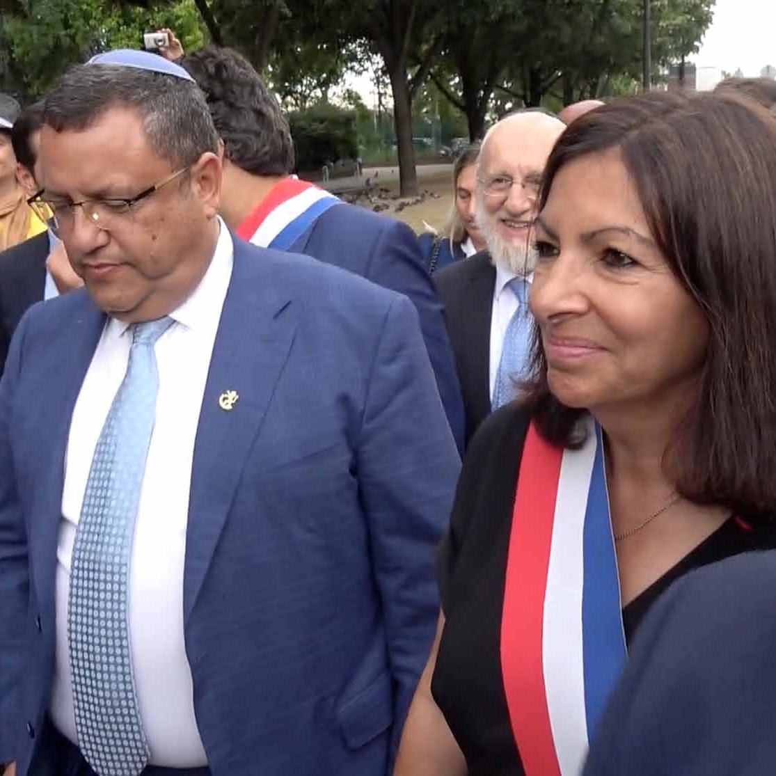 The image shows a group of people outdoors, likely at a public event. In the foreground, two individuals are prominently featured: one man is wearing a blue suit, and the other, a woman, is wearing a black outfit adorned with a ribbon that signifies a formal position. Both appear to be engaged in conversation or observing their surroundings. Other individuals are in the background, suggesting a social or official gathering. The setting appears to be a park or an outdoor area with trees.