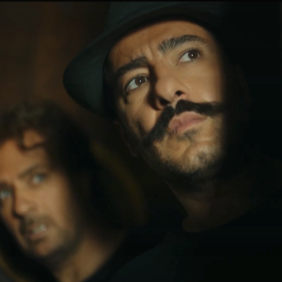 L'image montre deux hommes. L'un d'eux a une moustache prononcée et porte un chapeau, tandis que l'autre a les cheveux bouclés et semble inquiet. Ils regardent tous les deux vers la droite, comme s'ils étaient en train d'espionner ou de guetter quelque chose. L'éclairage est sombre, ce qui ajoute une atmosphère tendue et mystérieuse à la scène.