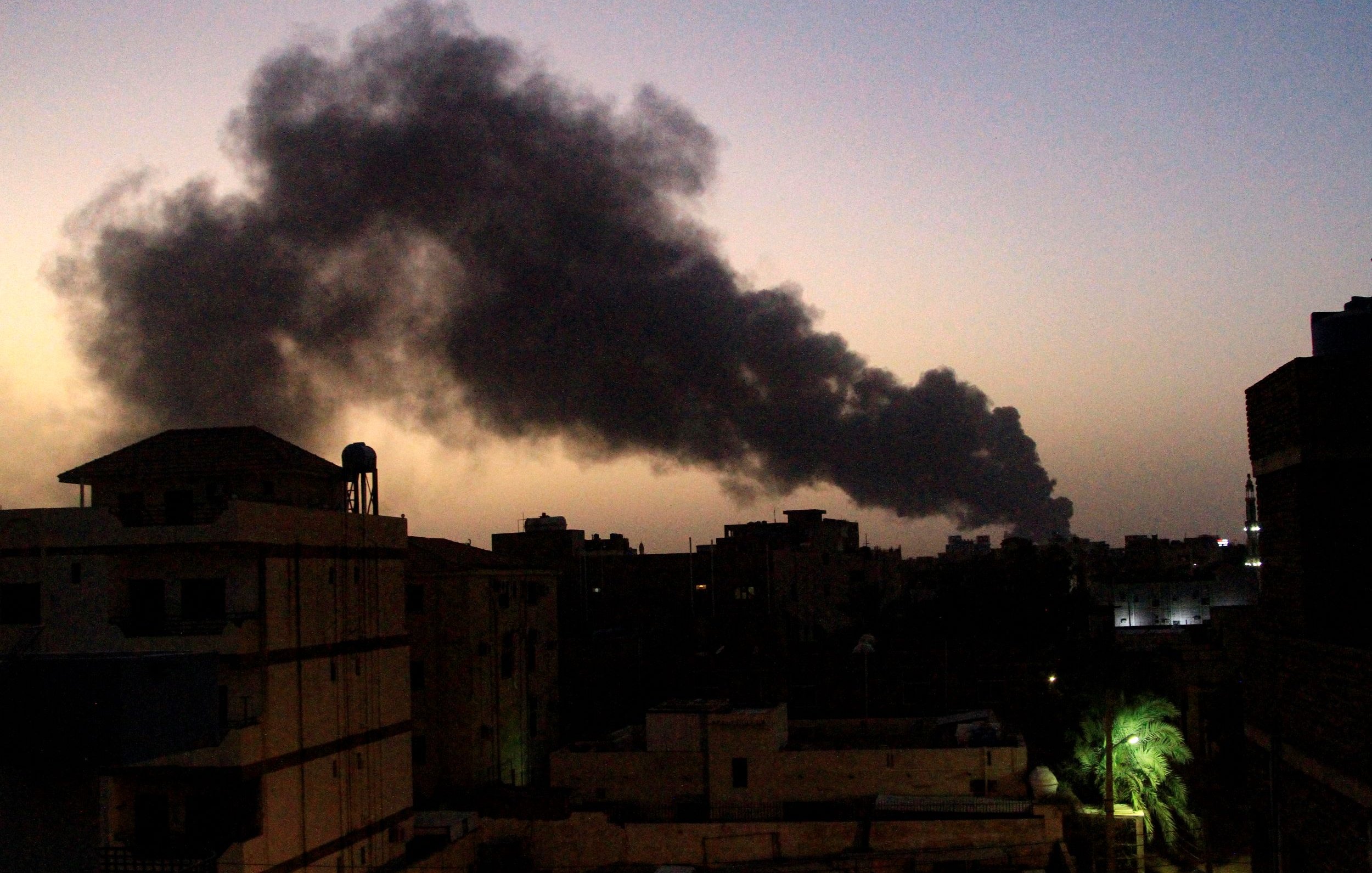 La imagen muestra un paisaje urbano al atardecer, con un cielo que presenta tonos oscuros. En el fondo, se puede ver una densa columna de humo negro que se eleva, sugiriendo la presencia de un incendio o alguna actividad industrial. A la izquierda, hay edificios que se recortan contra el cielo, y a la derecha, unas palmeras iluminadas por luces artificiales. La atmósfera parece tensa debido al humo y el entorno oscuro.