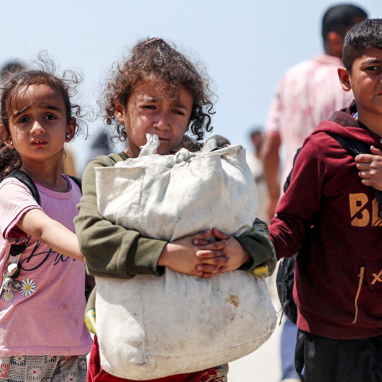 L'immagine mostra un gruppo di bambini che camminano, visibilmente in una situazione di difficoltà. Due bambine, una con i capelli ricci e l'altra con un'espressione preoccupata, accompagnano un ragazzo che tiene una grande borsa. Sembra che si trovino in un contesto di emergenza, con altre persone sullo sfondo. L'atmosfera è seriosa e trasmette una sensazione di vulnerabilità e speranza.