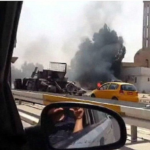 L'image montre une scène d'explosion ou d'incendie, avec de la fumée noire s'échappant d'un bâtiment en arrière-plan. On aperçoit également un véhicule jaune sur la route, tandis qu'une personne semble prendre la photo depuis l'intérieur d'une voiture. Des débris sont visibles près de l'endroit de l'incident, qui pourrait être lié à un conflit. La scène évoque une atmosphère de chaos et de tension.