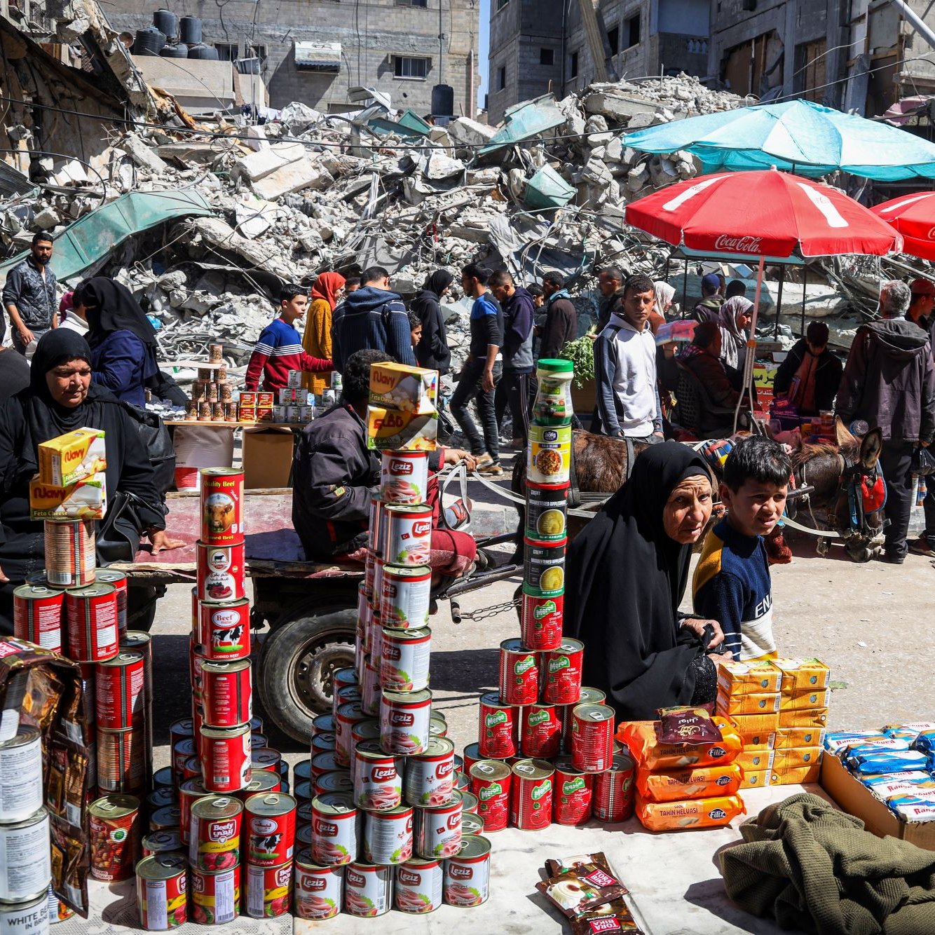 L'immagine mostra una scena di un mercato affollato, situato tra rovine di edifici. In primo piano, un gruppo di persone sta vendendo prodotti alimentari, con diversi ripiani di scatole e cibo in mostra. Sono visibili delle donne e dei bambini, alcuni seduti e altri in piedi, mentre diversi ombrelloni rossi forniscono ombra. Sullo sfondo, ci sono detriti di edifici crollati, che testimoniano una situazione di crisi. L'atmosfera sembra vivace nonostante le difficoltà evidenti.