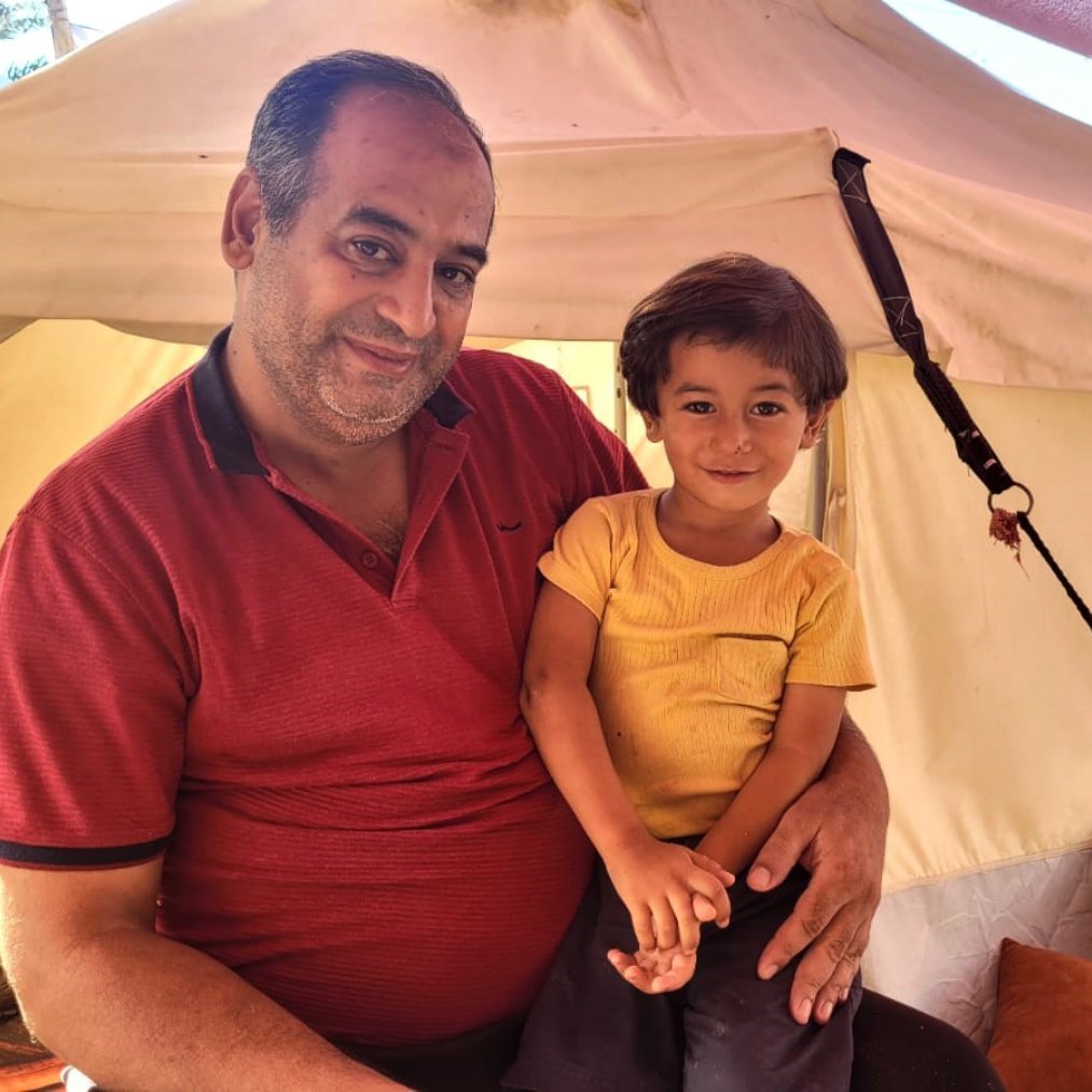 Nell'immagine si vede un uomo sorridente che tiene in braccio un bambino. L'uomo indossa una maglietta rossa e il bambino ha una maglietta gialla. Sono entrambi all'interno di una tenda, circondati da cuscini e altri oggetti. L'atmosfera sembra calda e accogliente, con una luce naturale che illumina la scena.