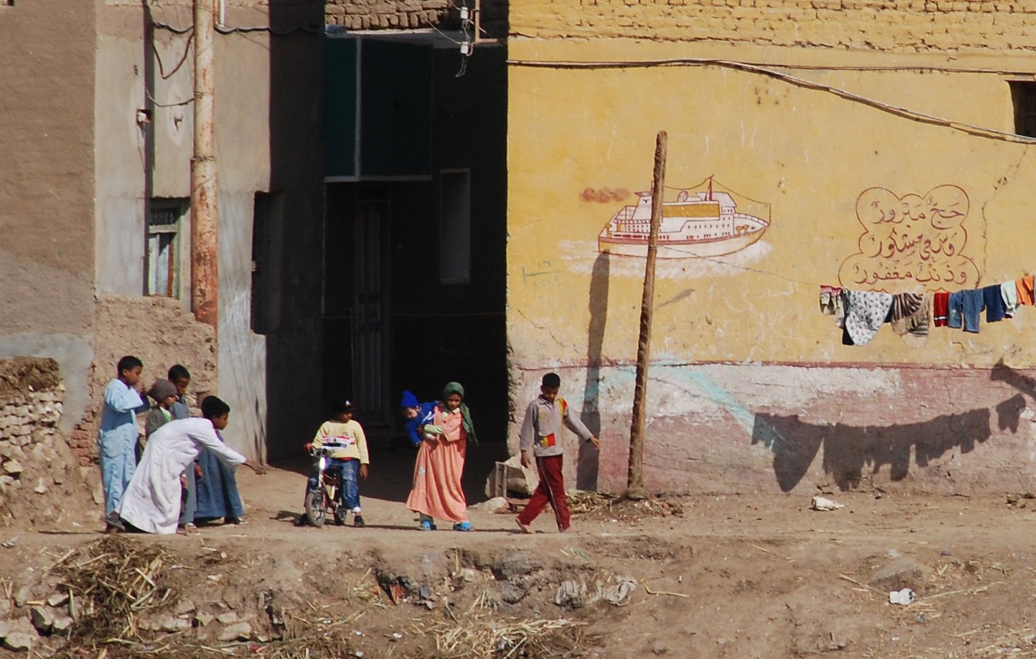 تظهر في الصورة مشهد لحياة يومية في منطقة شبه ريفية. في الجهة اليسرى يوجد شخص يرتدي ملابس بيضاء يحمل حقيبة، ويبدو أنه يسير نحو مجموعة من الأطفال والنساء. الأطفال يبدو أنهم يلعبون أو يتجهون إلى مكان ما. في الخلفية، توجد جدران ملونة تحتوي على رسوم جرافيتية وبعض الملابس المعلقة على حبل. الأرض تبدو جافة، مع بعض الحجارة والنباتات الصغيرة. يظهر كل ذلك الحياة البسيطة والمجتمع النشط في هذه المنطقة.