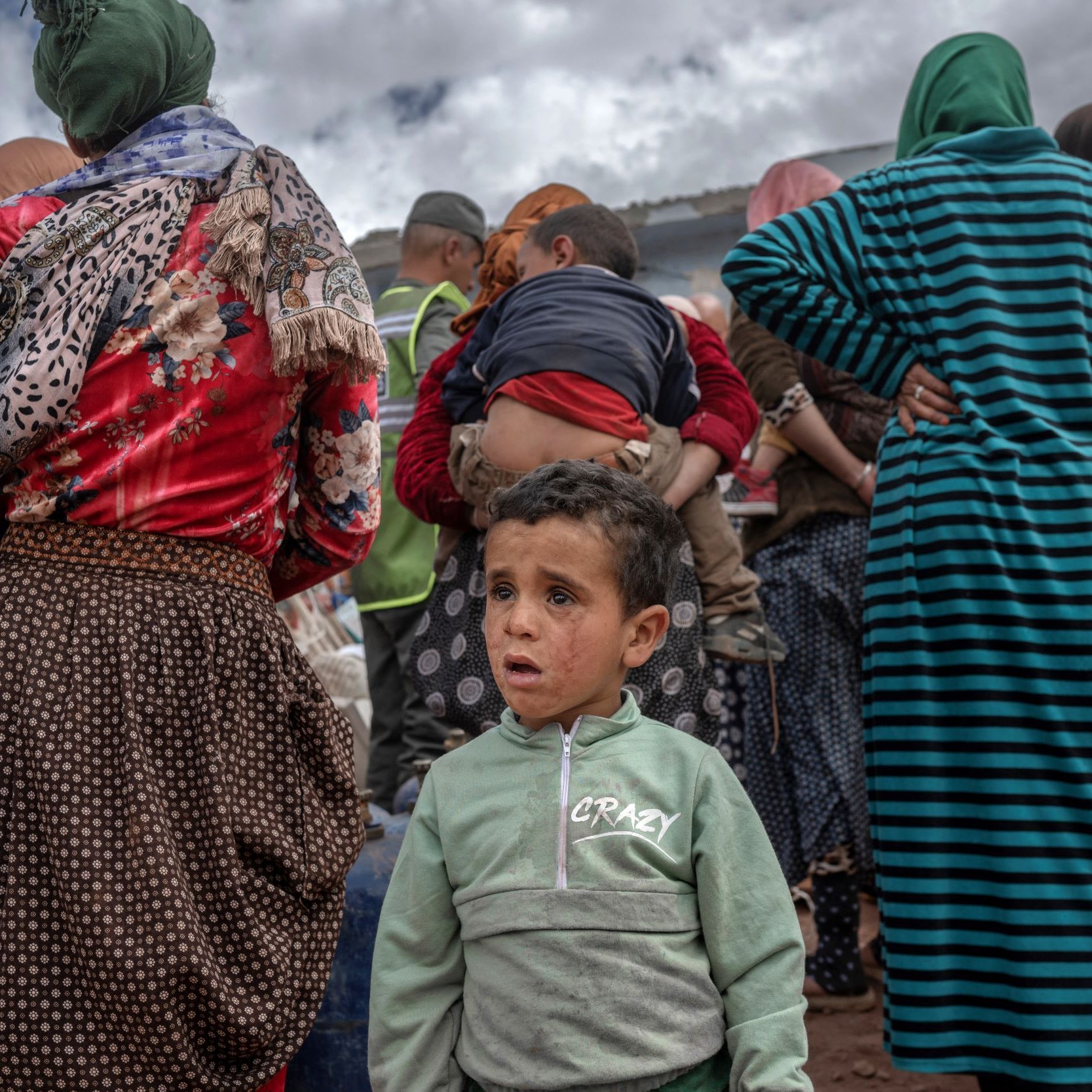 L'image montre un groupe de personnes, principalement des femmes en arrière-plan, portant des vêtements colorés et traditionnels. Parmi elles, un jeune garçon se tient au premier plan avec une expression préoccupée. Il porte une veste verte avec des écritures. Le décor suggère une atmosphère de rassemblement, peut-être dans un contexte de solidarité ou d'aide. Le ciel est nuageux, ajoutant une certaine gravité à la scène.