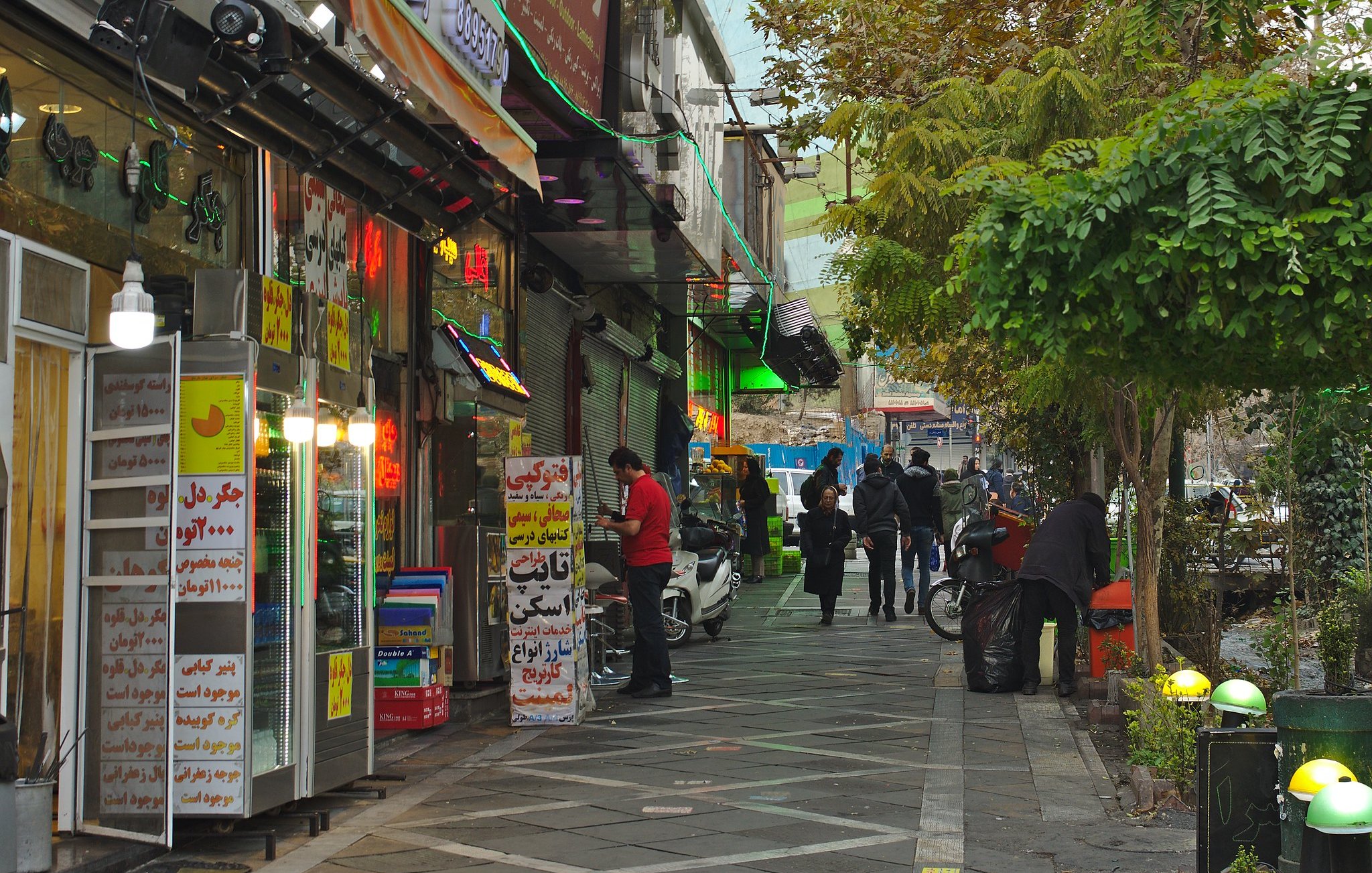 Une rue animée bordée de magasins colorés et de personnes marchant sous des arbres.