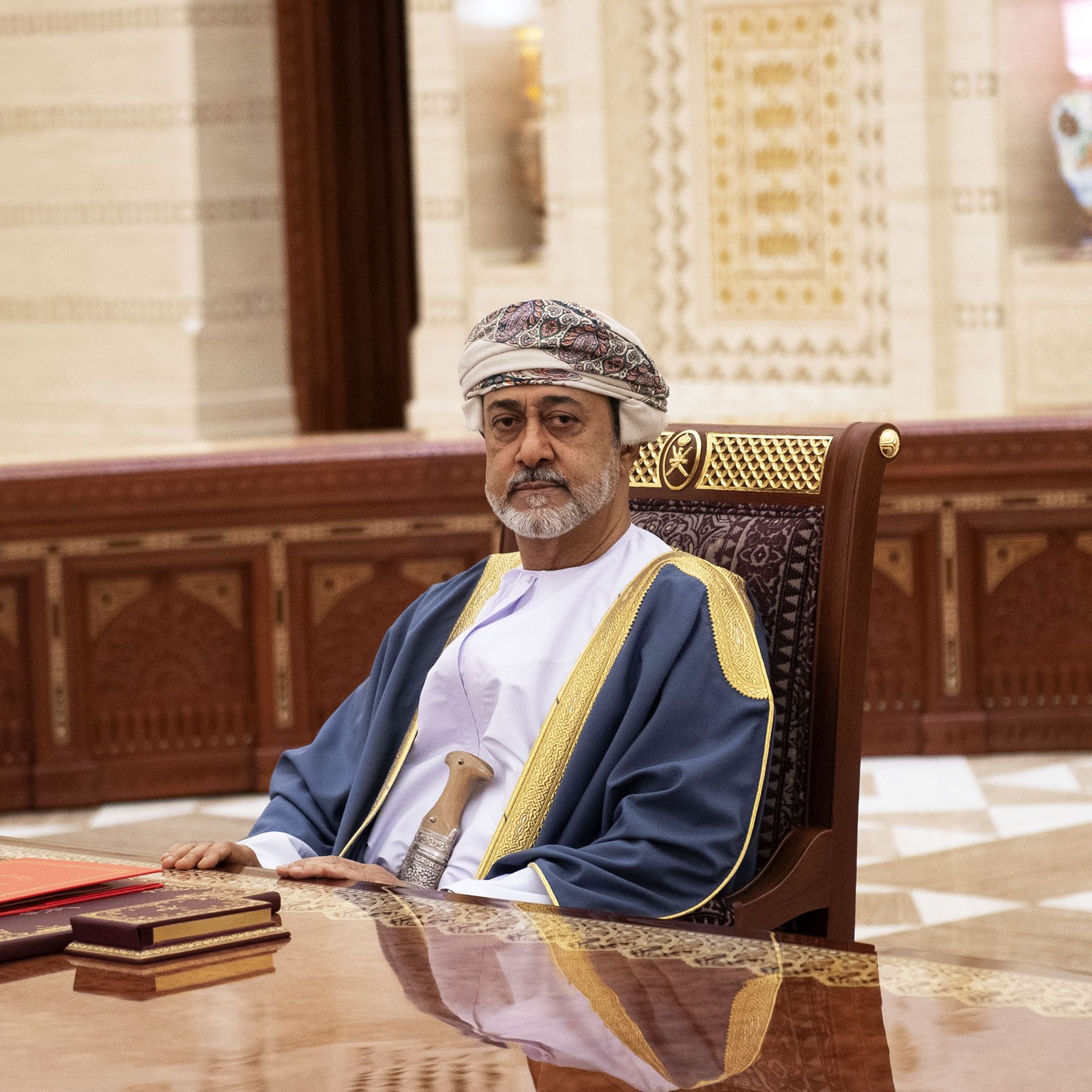 L'image montre un homme assis à un bureau dans un environnement intérieur richement décoré. Il porte un costume traditionnel, comprenant une robe longue et un turban orné. Le décor autour de lui présente des motifs architecturaux détaillés, typiques d'une ambiance élégante. Il semble concentré et sérieux, et le mobilier en bois ajoute à l'atmosphère formelle de la scène.