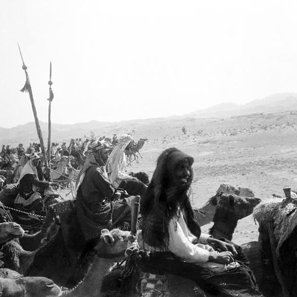 L'image montre un groupe de personnes montées sur des chameaux dans un paysage désertique. On peut voir des femmes et des hommes, certains portant des vêtements traditionnels. Au fond, des montagnes se dressent, et le ciel semble clair. Les personnes semblent participant à une sorte de caravane ou de procession, renforçant l'ambiance historique et culturelle de la scène.