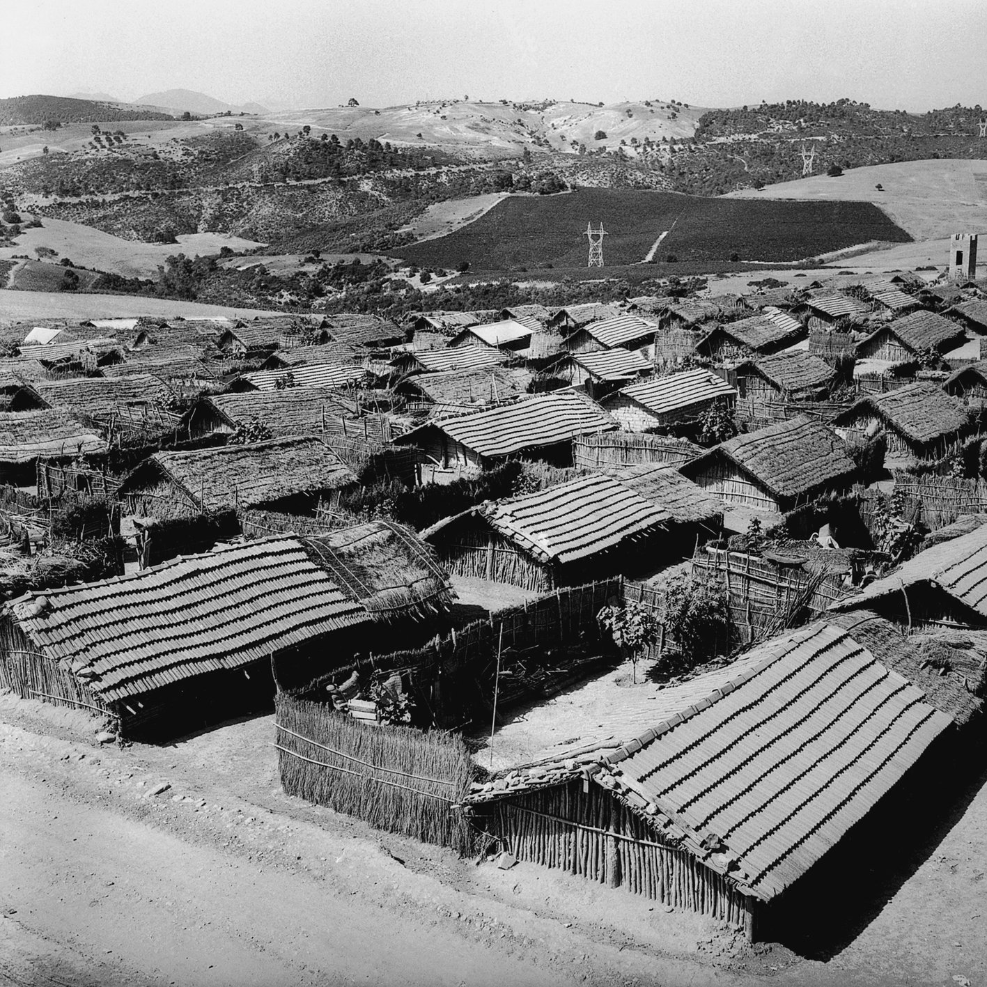 L'image montre un paysage rural avec plusieurs maisons en bois et en matériau naturel, probablement des huttes, disposées de manière dense. Les toits sont recouverts de paille ou d'un matériau similaire. On aperçoit également des collines verdoyantes à l'arrière-plan, ce qui suggère un environnement rural. Le ciel est clair, et l'ensemble de la scène a une tonalité noire et blanche, conférant une atmosphère nostalgique et ancienne à l'image.
