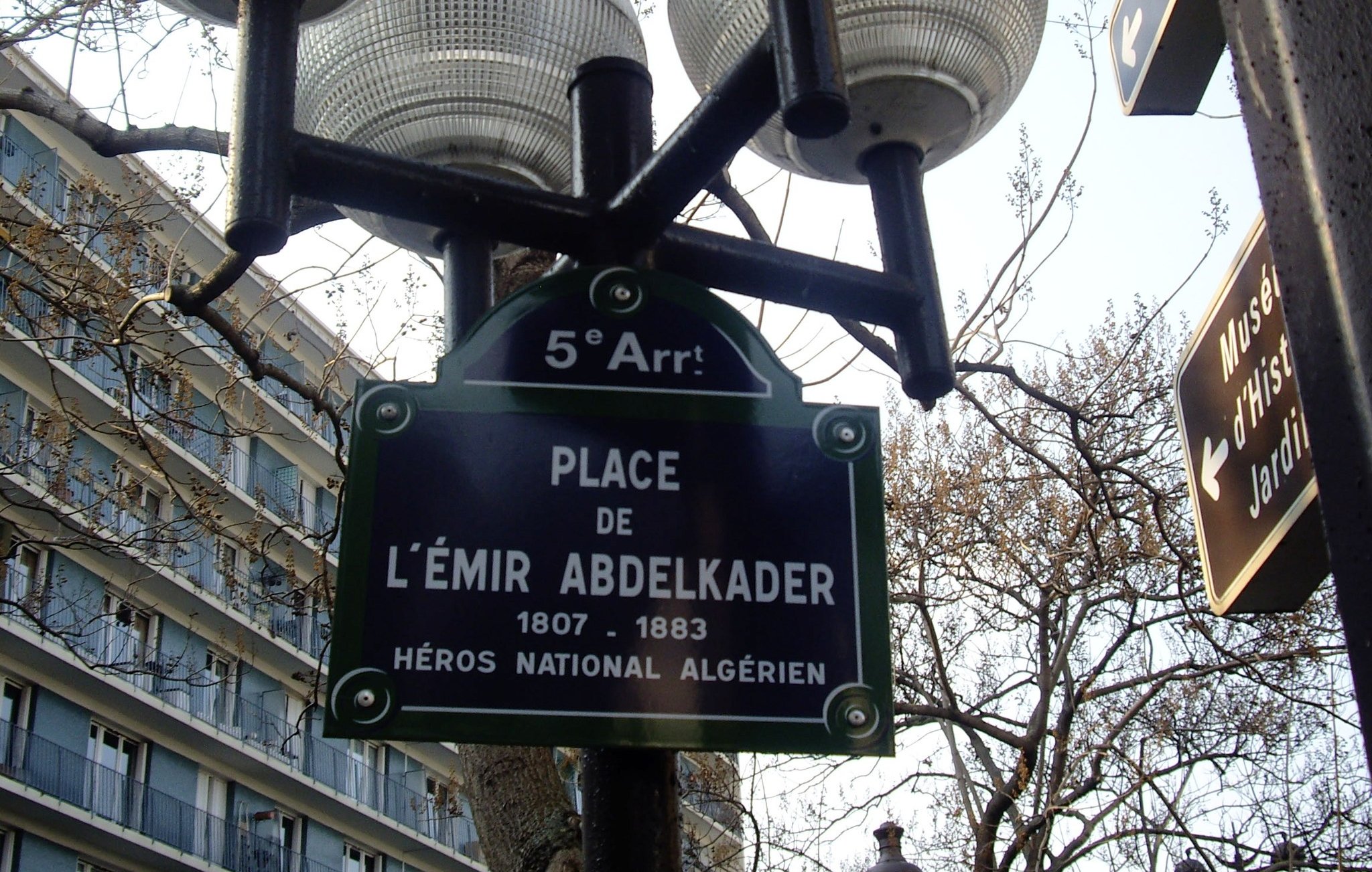 L'image montre un lampadaire avec plusieurs globes lumineux, sur lequel est fixée une plaque signalétique. Cette plaque indique "Place de l'Émir Abdelkader", accompagnée des dates 1807-1883 et de la mention "Héros National Algérien". Le tout se situe dans le cinquième arrondissement, comme le précise le petit panneau en haut. En arrière-plan, on peut apercevoir des bâtiments typiques de la ville.