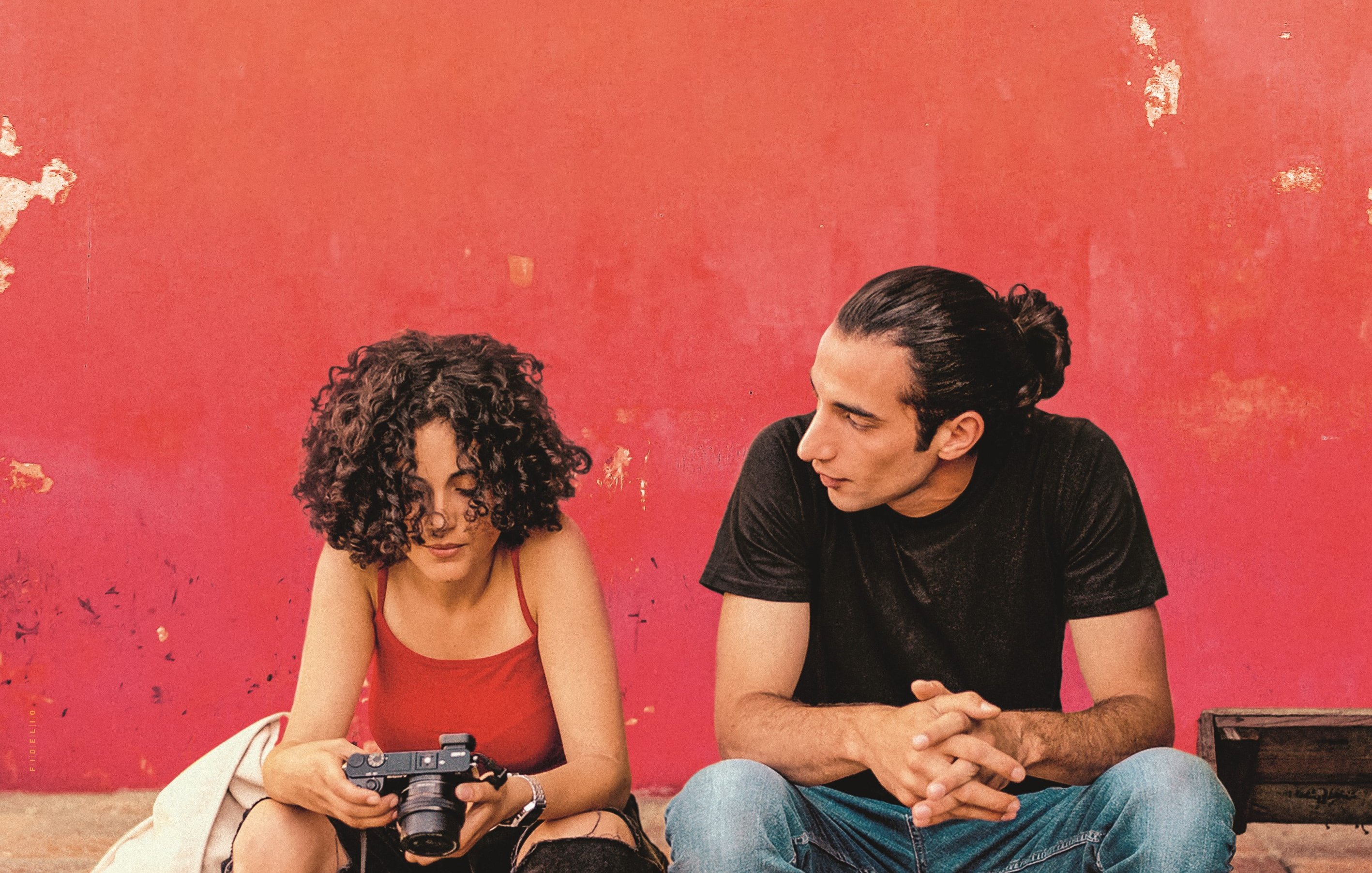 L'image montre deux personnes assises devant un mur rouge écaillé. La première personne, une femme aux cheveux bouclés, tient un appareil photo et semble concentrée sur son écran. Elle porte un débardeur rouge et un gilet clair. À côté d'elle, un homme avec des cheveux longs attachés en chignon la regarde attentivement. Il est vêtu d'un t-shirt noir et de jeans. L'atmosphère paraît détendue et intime, comme s'ils étaient en pleine conversation ou en train de partager un moment.