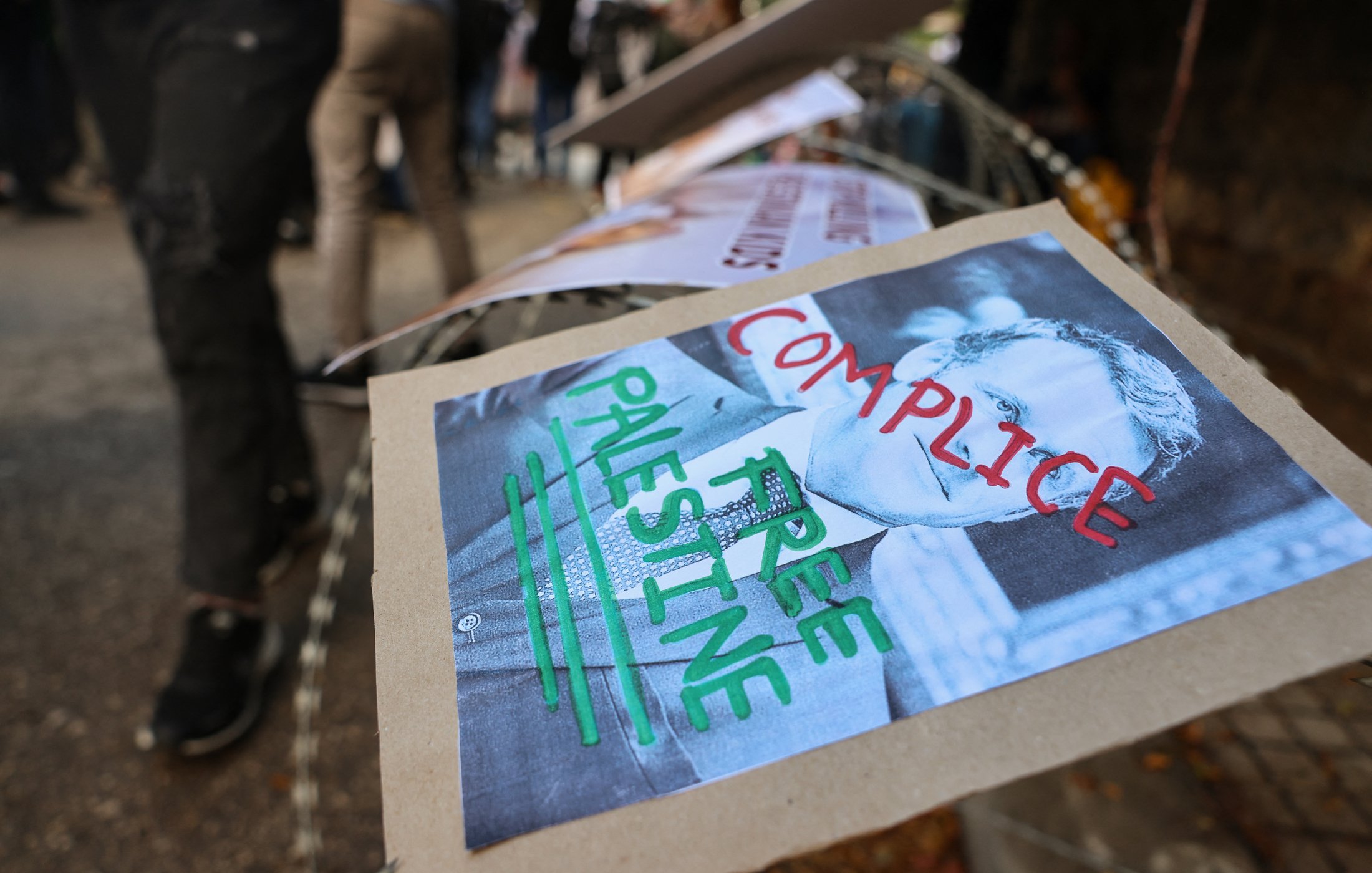 در تصویر، تعدادی از پوسترها روی یک نرده یا حصار قرار گرفته‌اند. یکی از این پوسترها تصویری از یک فرد همراه با نوشته‌هایی به زبان انگلیسی را نشان می‌دهد. نوشته‌های روی پوستر شامل کلماتی چون "COMPICILE" و "FREE PALESTINE" است. طراحی این پوستر به طور واضح به تضادهایی که بر سر مسائل فلسطین وجود دارد، اشاره دارد. زمینه‌ای غیررسمی و معترضانه به نظر می‌رسد و نشانه‌های حمایت از فلسطین را به تصویر می‌کشد.