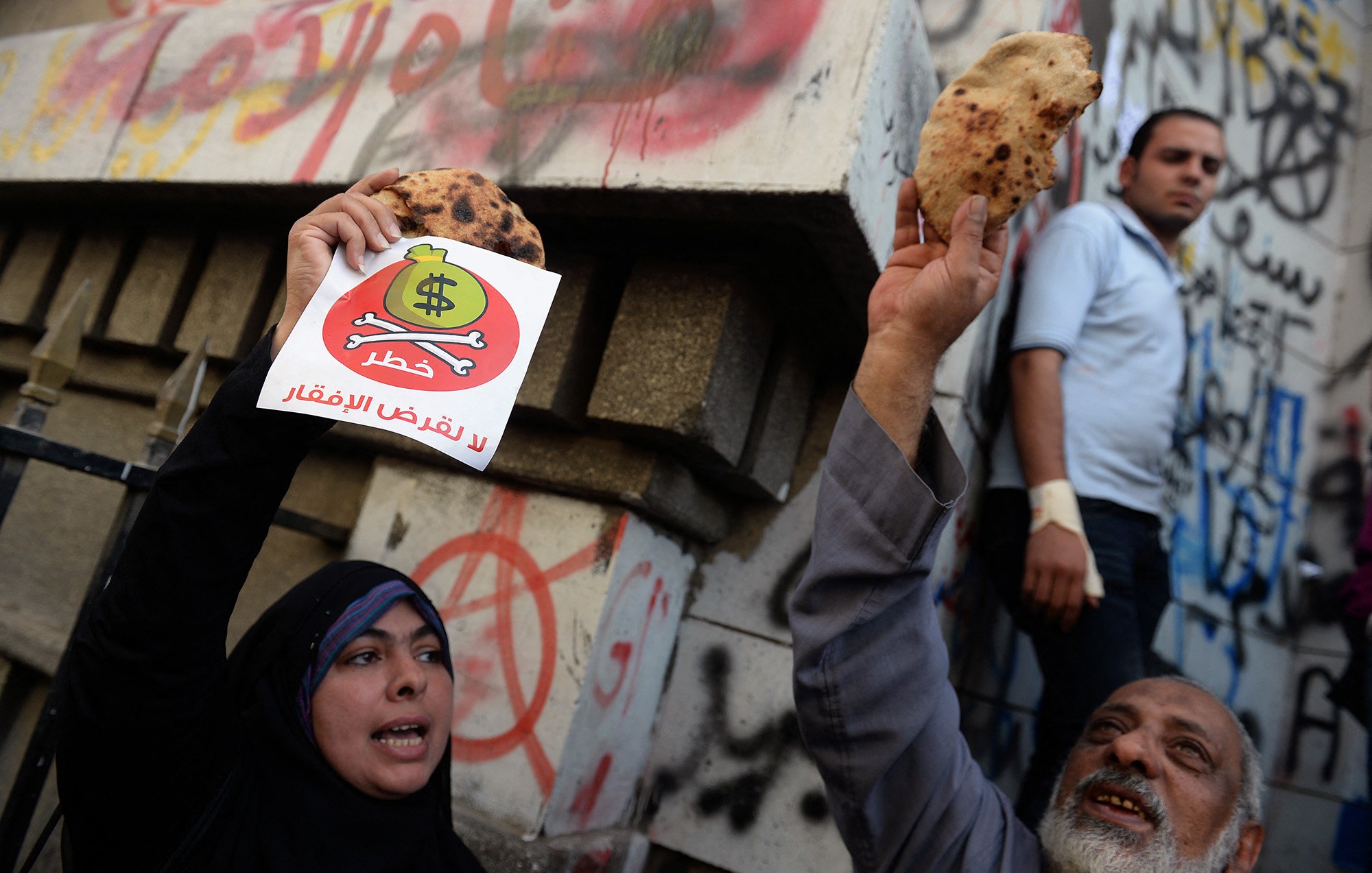 الصورة تظهر مجموعة من الأشخاص في تظاهرة. في المقدمة، يوجد رجل وامرأة يحملان لافتات. المرأة ترتدي الحجاب وتبدي حماسًا بالرفع لافتة مكتوب عليها شعار، بينما الرجل يحمل قطعة خبز. في الخلفية، تظهر جدران مكتوبة عليها شعارات وشعارات فنية. يبدو أن المتظاهرين يعبرون عن رأيهم حول موضوع معين، وملابسهم تشير إلى أجواء الاحتجاج.