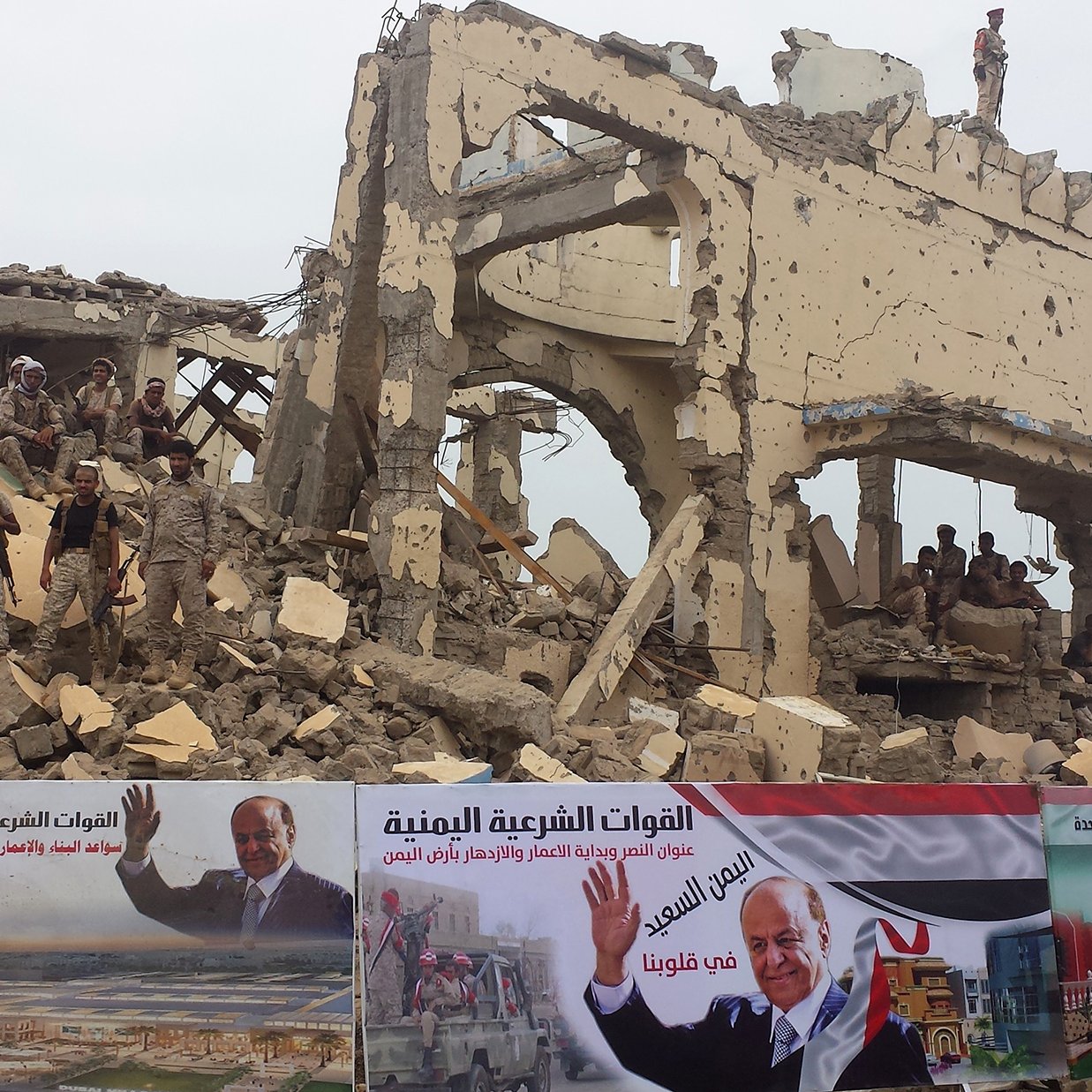 The image depicts a heavily damaged building that has partially collapsed, suggesting that it may be the result of conflict or disaster. In the foreground, there are several large banners featuring messages and images, including one that appears to be a political endorsement. Soldiers are visible on top of the rubble, indicating a military presence in the area. The overall scene conveys a sense of destruction and the impact of ongoing turmoil, likely in a war-affected region.