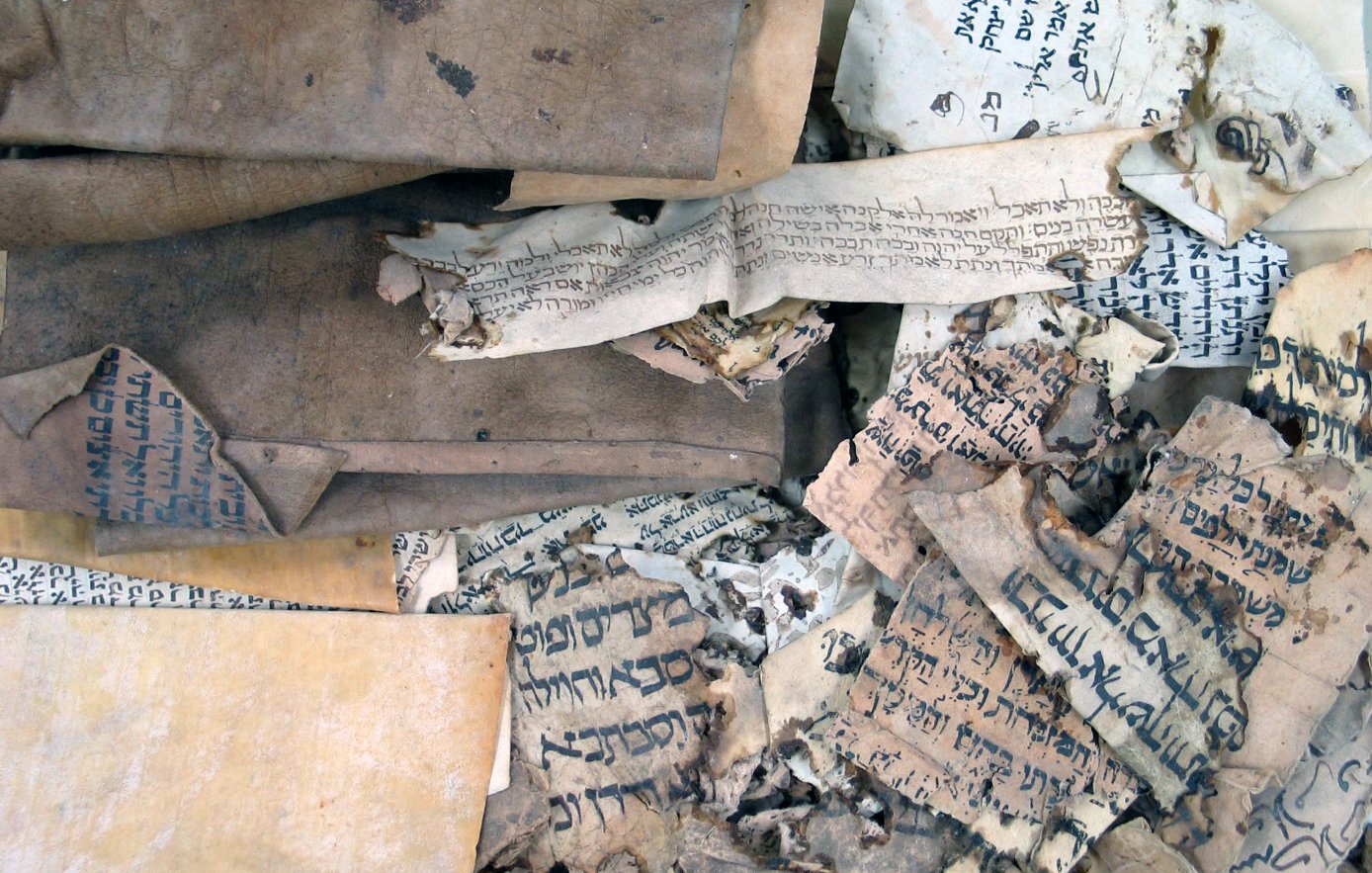 L'image montre un ensemble de vieux manuscrits et de documents enroulés et usés. Les pages sont usées, certaines sont jaunies et déchirées, et on peut voir des inscriptions écrites dans des langues anciennes et probablement des alphabets différents. La texture des papiers varie, certains étant plus épais que d'autres, et la composition générale donne une impression d'antiquité et de fragilité, suggérant leur importance historique.