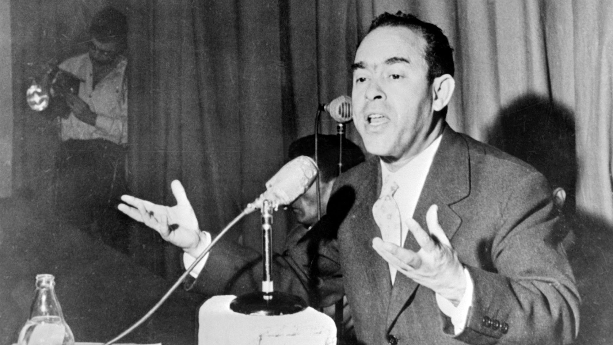 L'image montre un homme en train de s'exprimer avec enthousiasme lors d'une conférence ou d'un discours. Il est vêtu d'un costume et parle devant un microphone, gesticulant avec les mains pour accentuer ses propos. Sur la table devant lui, on peut voir un bloc de béton et quelques documents. En arrière-plan, une autre personne semble prendre des photos ou enregistrer. L'ambiance paraît sérieuse, soulignant l'importance de son discours.