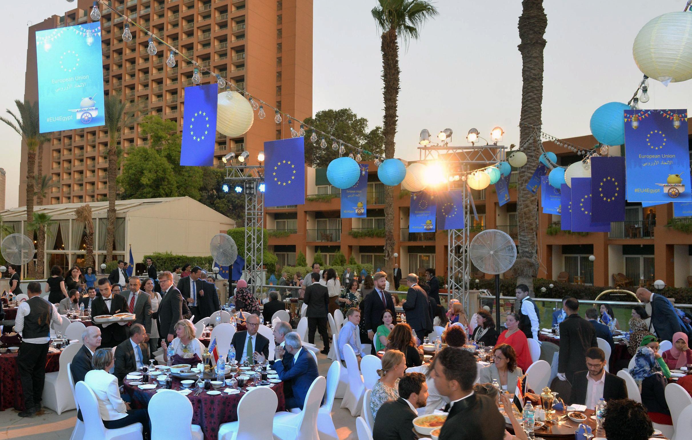 L'image montre un événement extérieur festif, probablement un dîner ou une réception. Des tables rondes sont disposées avec des nappes élégantes, où des convives en tenues formelles s'assoient pour profiter du repas. L'environnement est décoré avec des lanternes et des bannières aux couleurs de l'Union européenne. Le décor comprend également des palmiers et des installations lumineuses, créant une ambiance conviviale et festive au crépuscule. Les personnes présentes semblent converser joyeusement et profiter de l'événement.
