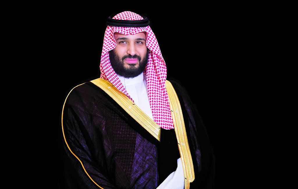 L'image montre un homme vêtu d'une tenue traditionnelle saoudienne, composée d'une darra'a noire avec des ornements dorés et d'un keffieh rouge et blanc, maintenu par une agrafe noire. Il affiche une expression sérieuse et confiante, se tenant droit avec les bras légèrement croisés devant lui. Le fond est sombre, ce qui met en valeur sa tenue et son apparence.