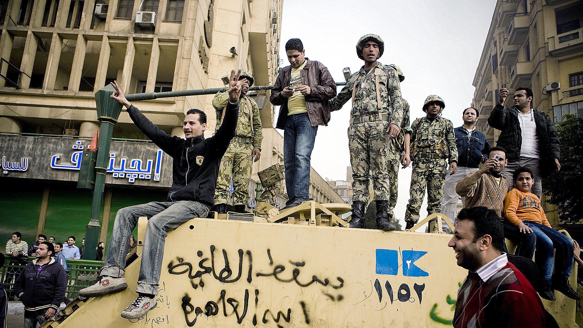 در این تصویر، یک گروه از مردان و سربازان در حال تجمع روی یک خودرو نظامی هستند. چند نفر از آنها به سمت دوربین خوشحالی می‌کنند و دست بلند کرده‌اند. در پس‌زمینه، ساختمان‌هایی با نشانه‌ها و نوشته‌هایی به زبان عربی دیده می‌شود. فضای تصویر نشان‌دهنده یک وضعیت اجتماعی یا سیاسی است که در آن افراد در حال ابراز احساسات و تعامل با یکدیگر هستند.