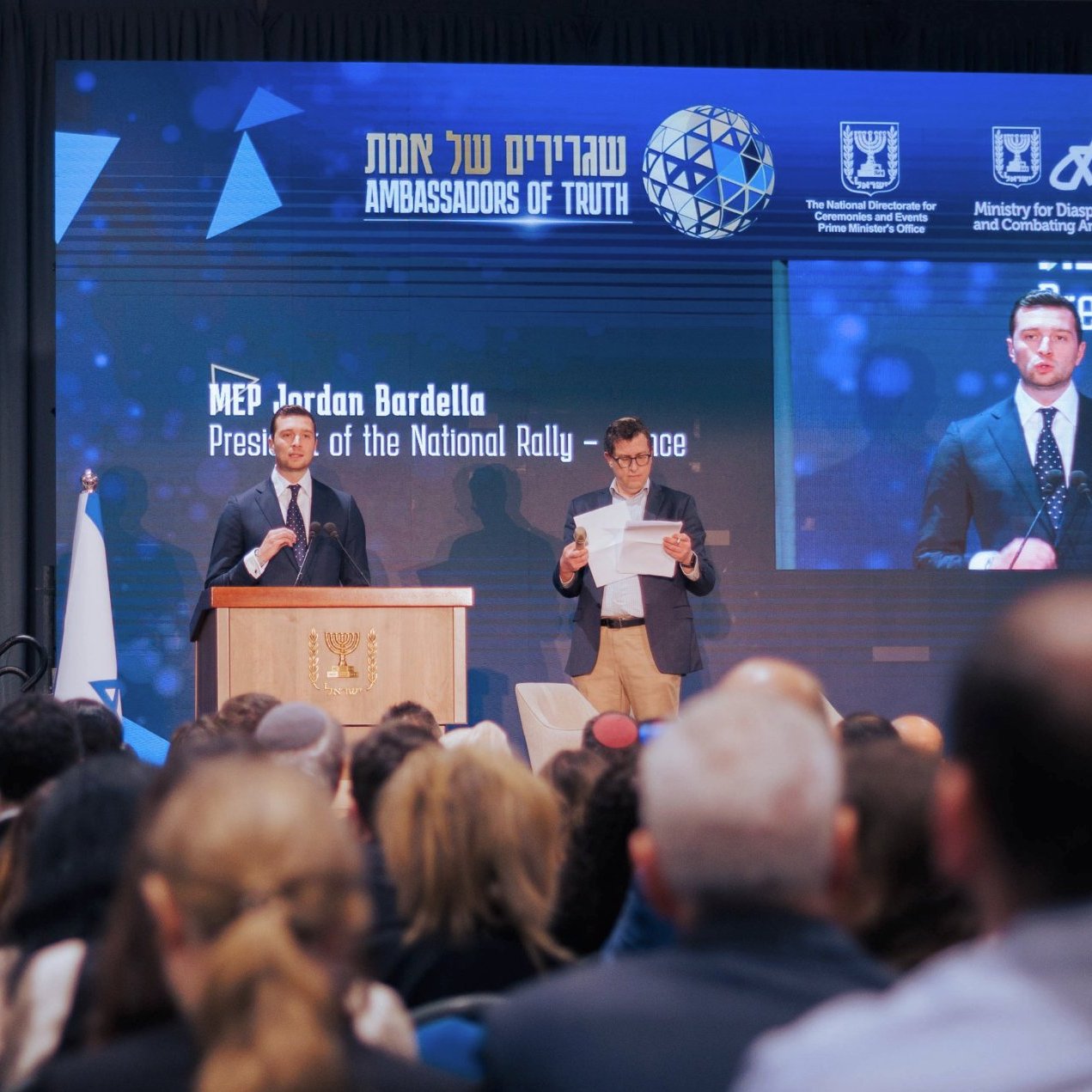 الصورة تظهر حدثًا رسميًا حيث يتحدث متحدث أمام جمهور كبير. على المنصة، يوجد شخص يحمل ورقة، ويبدو أن المتحدث يقرأ من نص. هناك خلفية تعرض شعارًا ومعلومات عن الفعالية. الأجواء تبدو جدية والر audience مهتم.