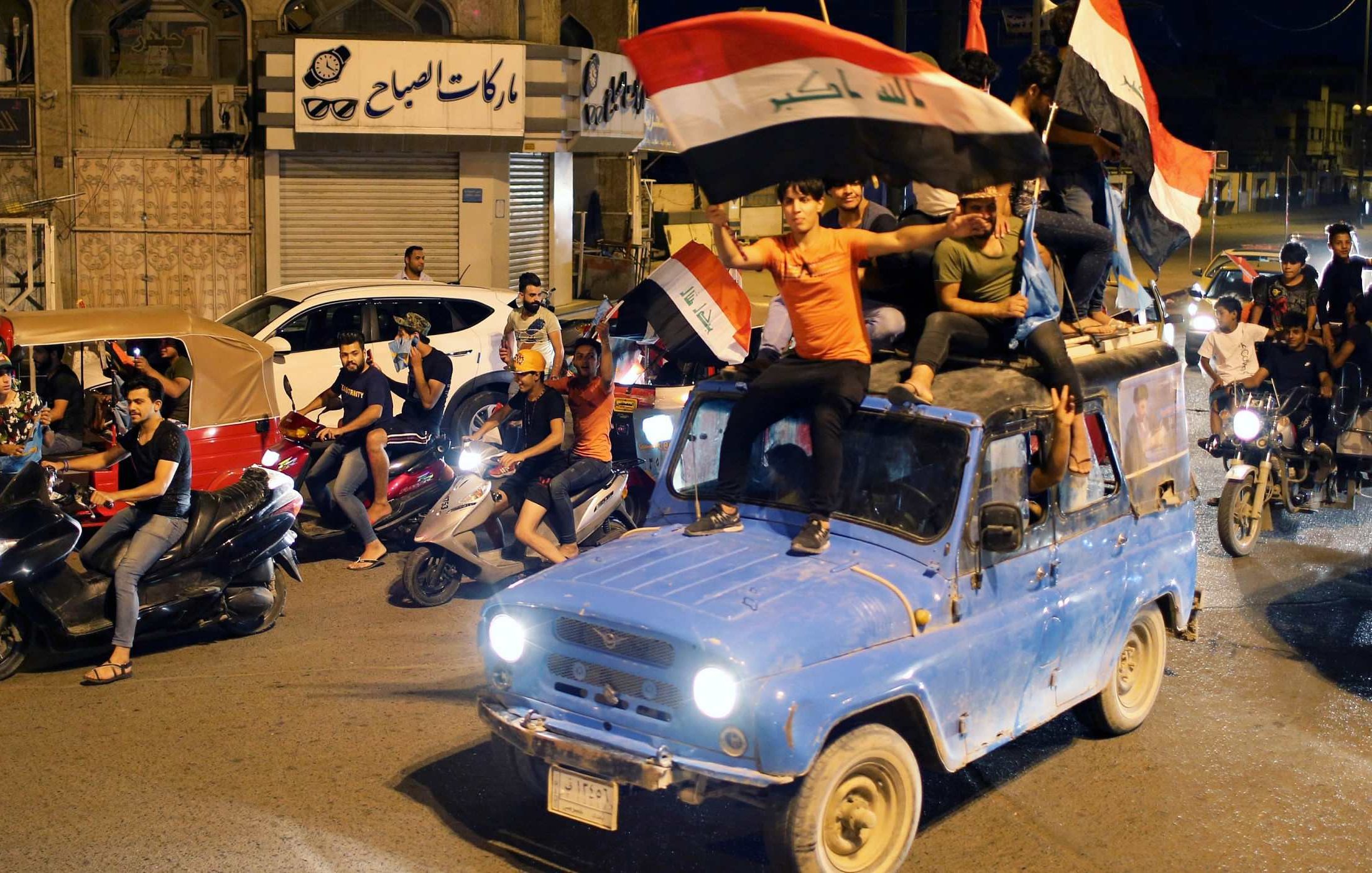 L'image représente une scène de célébration dans une rue animée. Des personnes sont rassemblées autour de véhicules, certains montent sur un ancien pickup bleu. Des drapeaux irakiens sont agités, tandis que d'autres participants conduisent des motos et des scooters. L'atmosphère est festive et dynamique, indiquant un moment de joie collective. Des bâtiments sont visibles en arrière-plan, suggérant une ambiance urbaine.