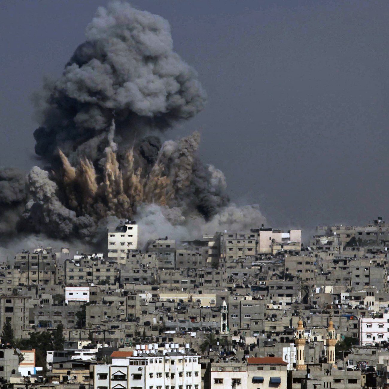 Nell'immagine si vede un'esplosione che solleva una grande nuvola di fumo grigio scuro sopra una città. Gli edifici della città sono visibili in primo piano, molti dei quali appaiono danneggiati o distrutti. La scena evoca un senso di devastazione e conflitto, con il cielo che si oscura a causa del fumo e dei detriti sollevati dall'esplosione.