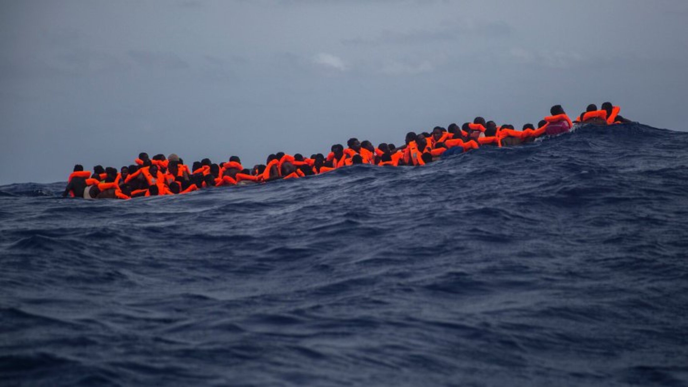 L'image montre un groupe de personnes en mer, portant des gilets de sauvetage orange. Elles semblent rassemblées sur une petite embarcation, luttant contre des vagues dans une étendue d'eau bleue profonde. Le ciel est nuageux, ce qui accentue la gravité de la situation maritime. Cette scène évoque des thèmes de migration, de survie et de danger en mer.