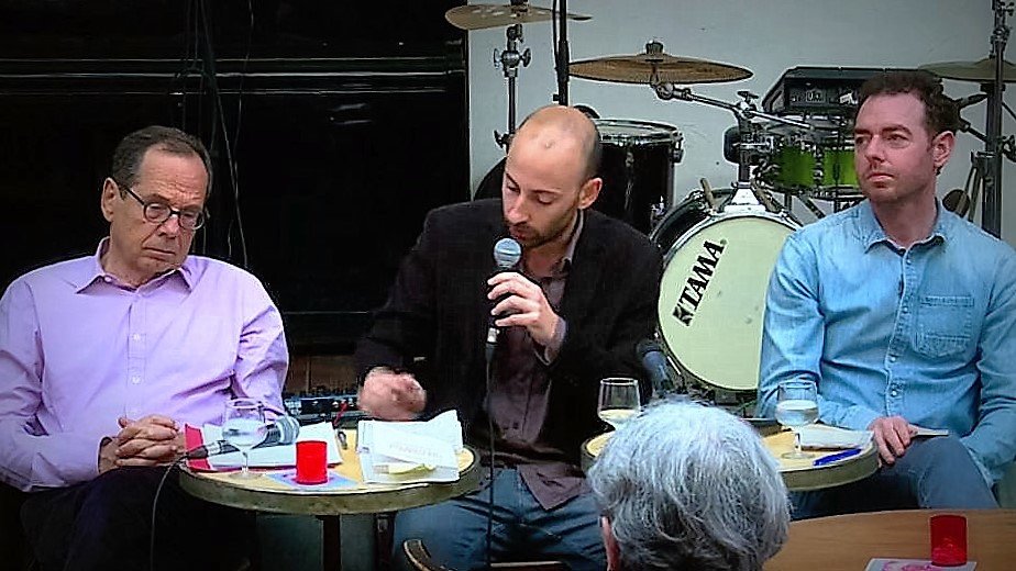 L'image montre un moment lors d'un événement où trois hommes sont assis autour de petites tables. Au centre, un homme parle dans un micro, semblant engager une discussion ou une présentation. Les autres hommes, à sa gauche et à sa droite, écoutent attentivement. On peut voir des instruments de musique en arrière-plan, ce qui suggère que l'événement se déroule dans un cadre décontracté, peut-être un café ou un bar. Des verres et des notes sont présents sur les tables, ajoutant à l'ambiance informelle de la rencontre.