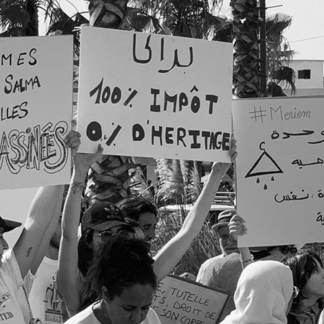 L'image est en noir et blanc et montre un rassemblement de personnes tenant des pancartes. Certaines des pancartes portent des messages en français comme : - "Nous sommes Aïcha, Samiha, Salma et toutes celles qu'ils ont ASSASSINÉES" - "100% IMPÔT 0% D'HÉRITAGE" Une autre pancarte comporte des inscriptions en arabe et le hashtag "#Meviem." Les personnes dans l'image semblent être engagées dans une manifestation ou une protestation concernant les droits des femmes et les questions de violence ou d'injustice. On peut également voir quelques palmiers et des bâtiments en arrière-plan, ce qui suggère que la scène se déroule en plein air dans un lieu urbain.