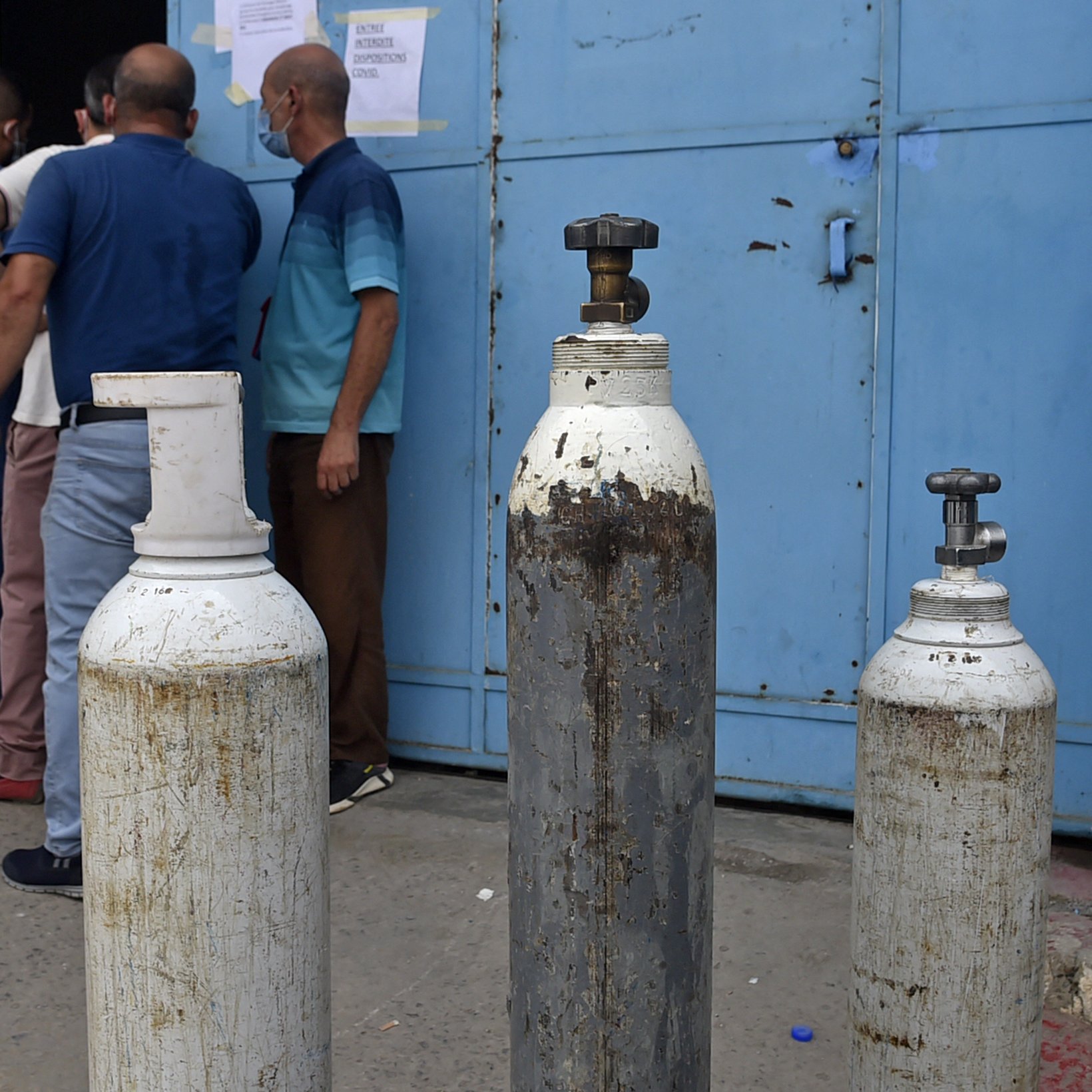L'image montre un groupe de personnes devant une porte bleue. Sur le devant, il y a plusieurs bouteilles de gaz, dont certaines sont blanches et d'autres en métal. Les bouteilles semblent usées et peuvent être utilisées pour stocker des gaz, comme l'oxygène. Les personnes à l'arrière semblent attendre ou discuter, tandis que l'environnement semble être un lieu urbain.