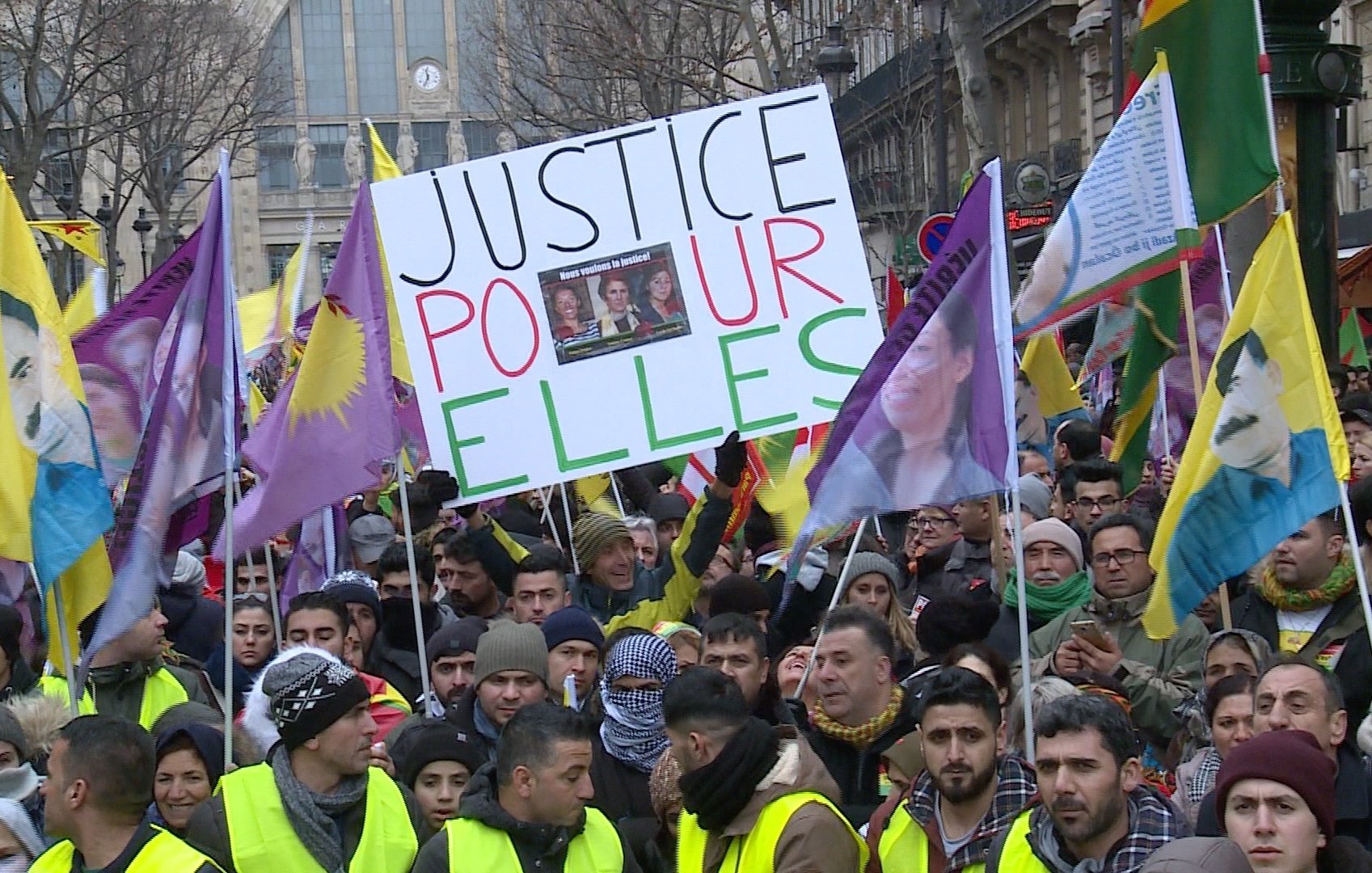 L'image montre une manifestation avec de nombreuses personnes rassemblées. Les manifestants portent des drapeaux colorés et des panneaux. L'un des panneaux affiche le message "JUSTICE POUR ELLES", suggérant un appel à la justice, probablement pour des femmes. Les visages des manifestants sont déterminés, et l'atmosphère semble impliquée et engagée.