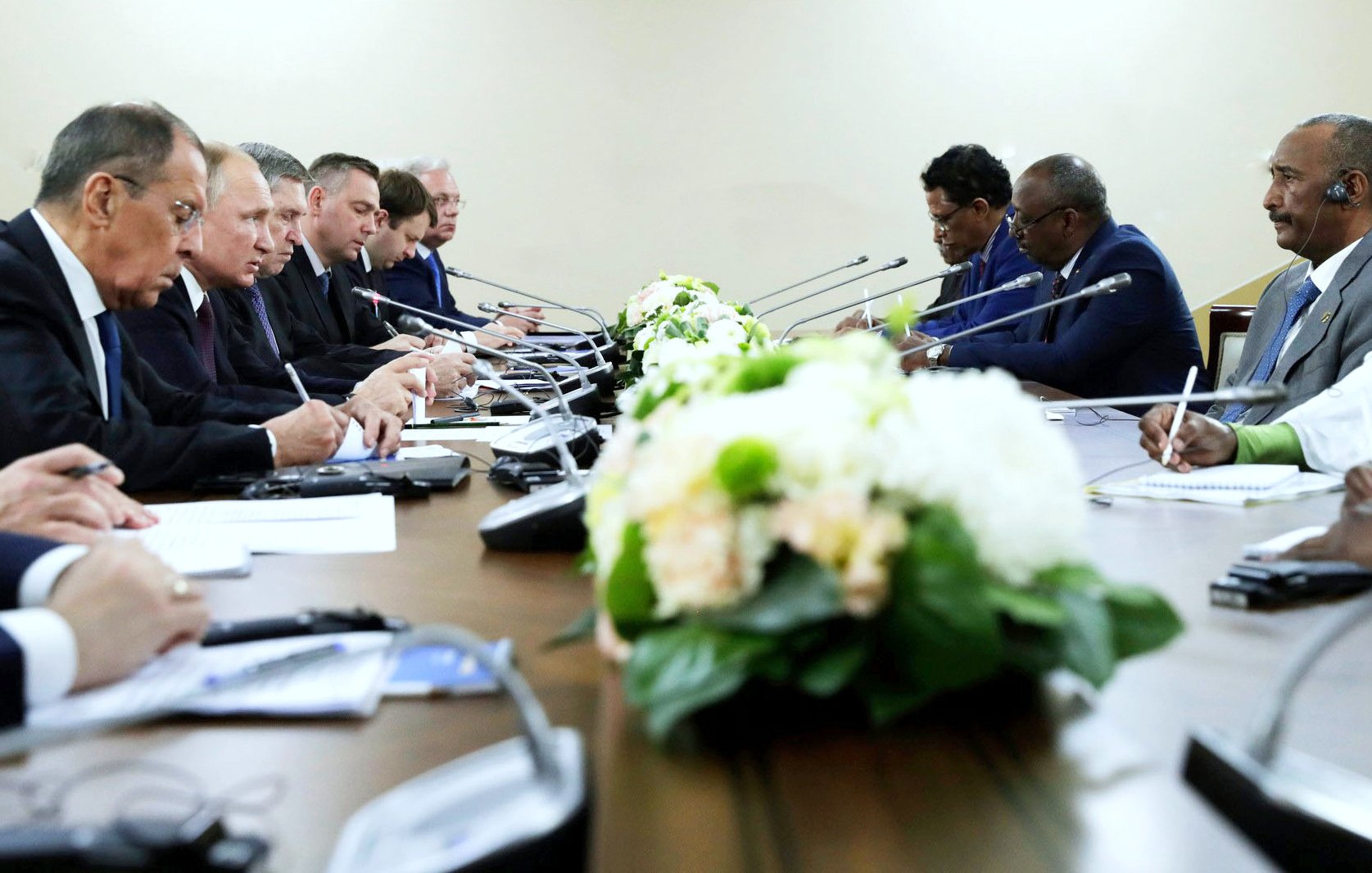 La imagen muestra una reunión formal en una sala con una mesa larga. Hay varias personas sentadas a ambos lados de la mesa, concentradas en la conversación. En el centro de la mesa, hay un arreglo floral decorativo. Las personas parecen estar intercambiando ideas, y algunos están tomando notas. El ambiente es serio y profesional.