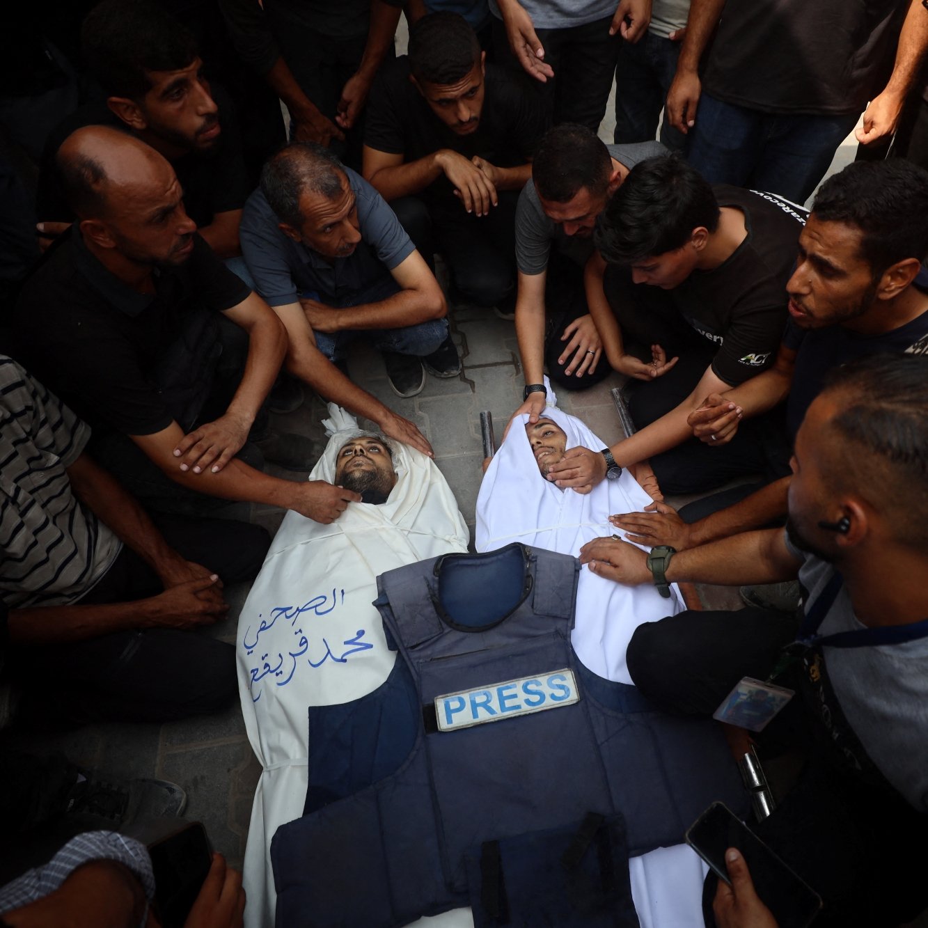 A crowd mourns over a deceased person covered in a white cloth, wearing a press vest.
