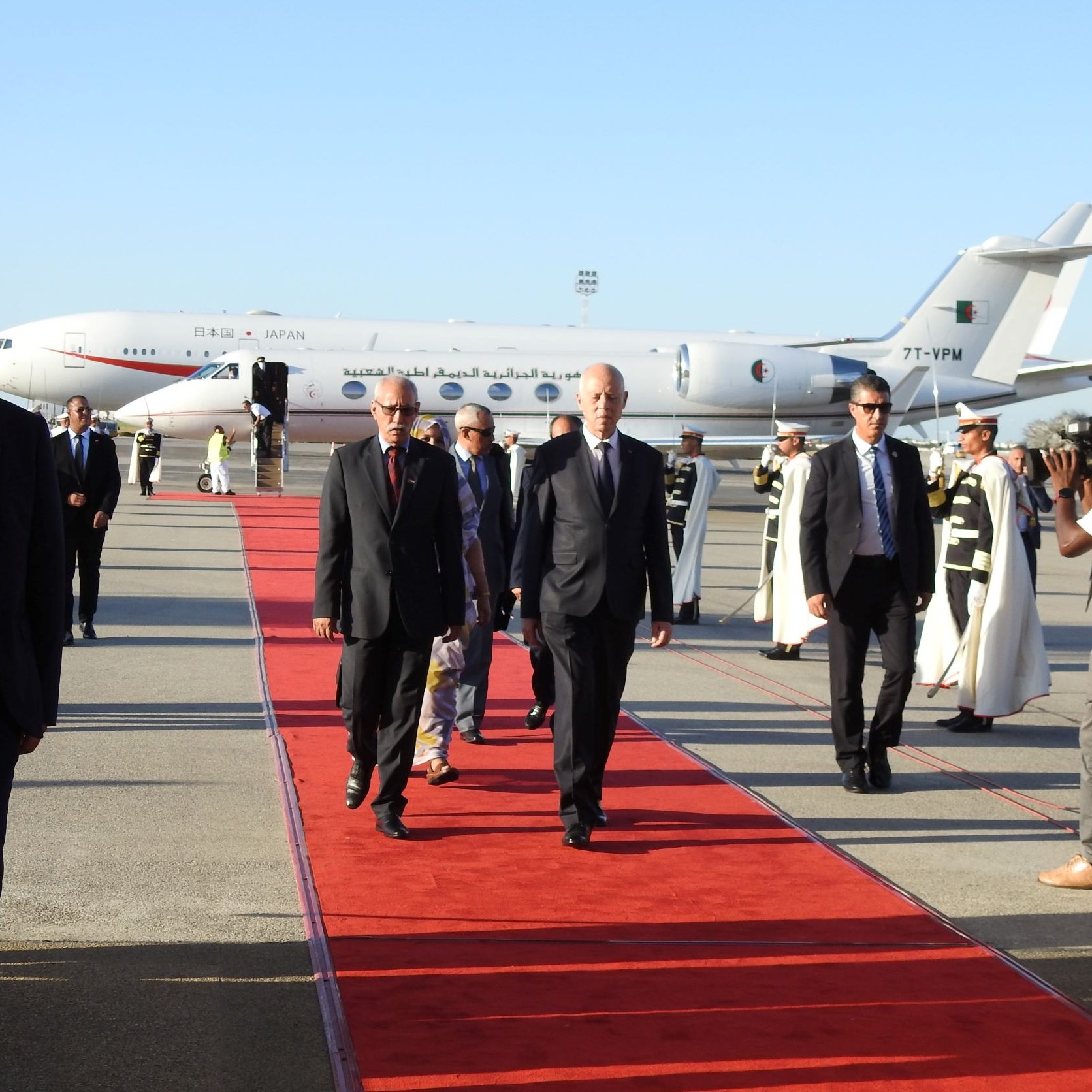 L'image montre un aéroport avec une piste. Un avion, visible en arrière-plan, est stationné. Des personnes marchent sur un tapis rouge, probablement des dignitaires ou des personnalités importantes. Certains hommes en costume accompagnent les personnes sur le tapis, tandis que d'autres, habillés en tenue formelle, semblent filmer ou prendre des photos de l'événement. L'atmosphère est formelle et festive, indiquant une arrivée importante.